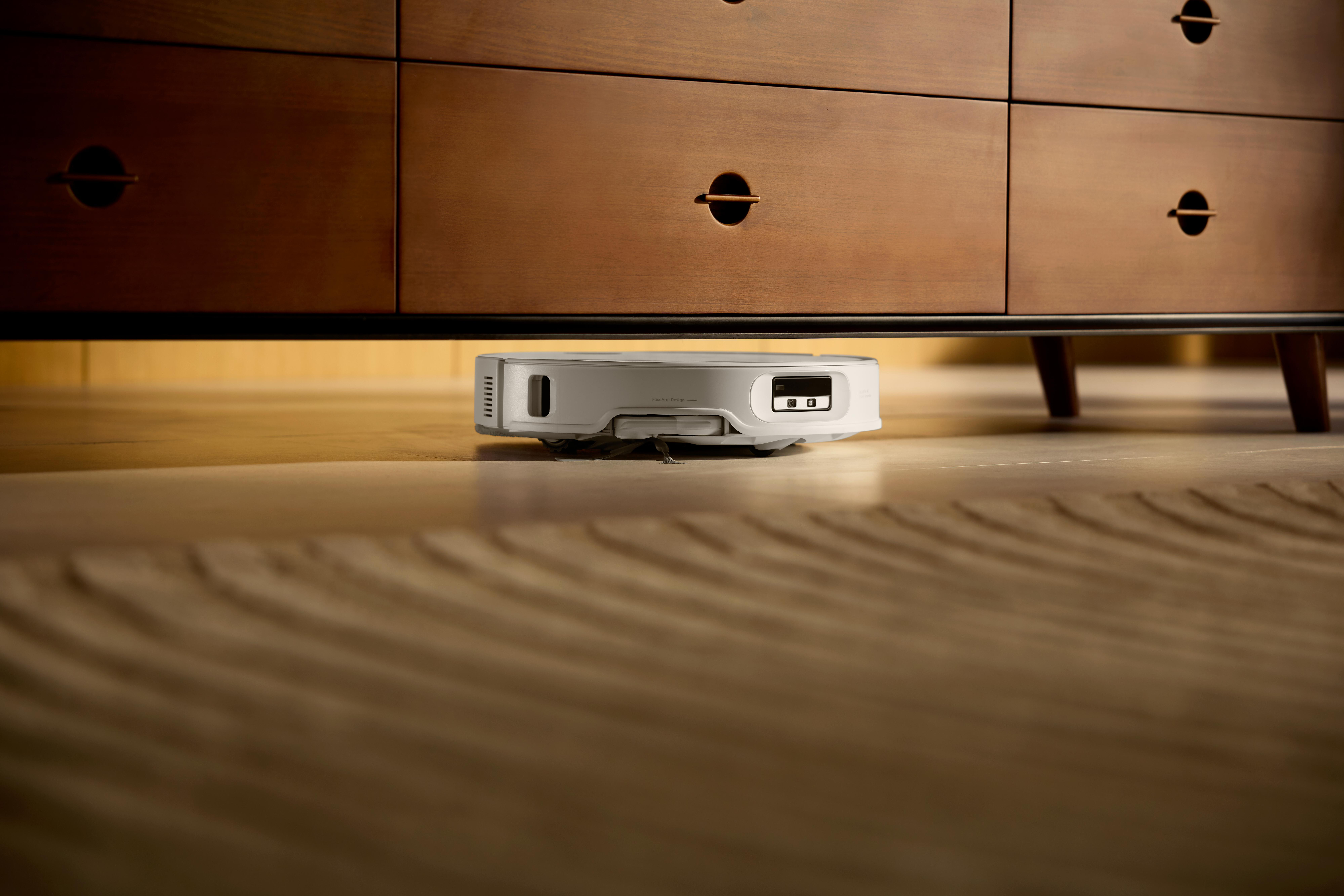 Ein Roboter-Staubsauger unter einem Holzschrank. Der Schrank hat eine dunkelbraune Oberfläche und runde Griffe.