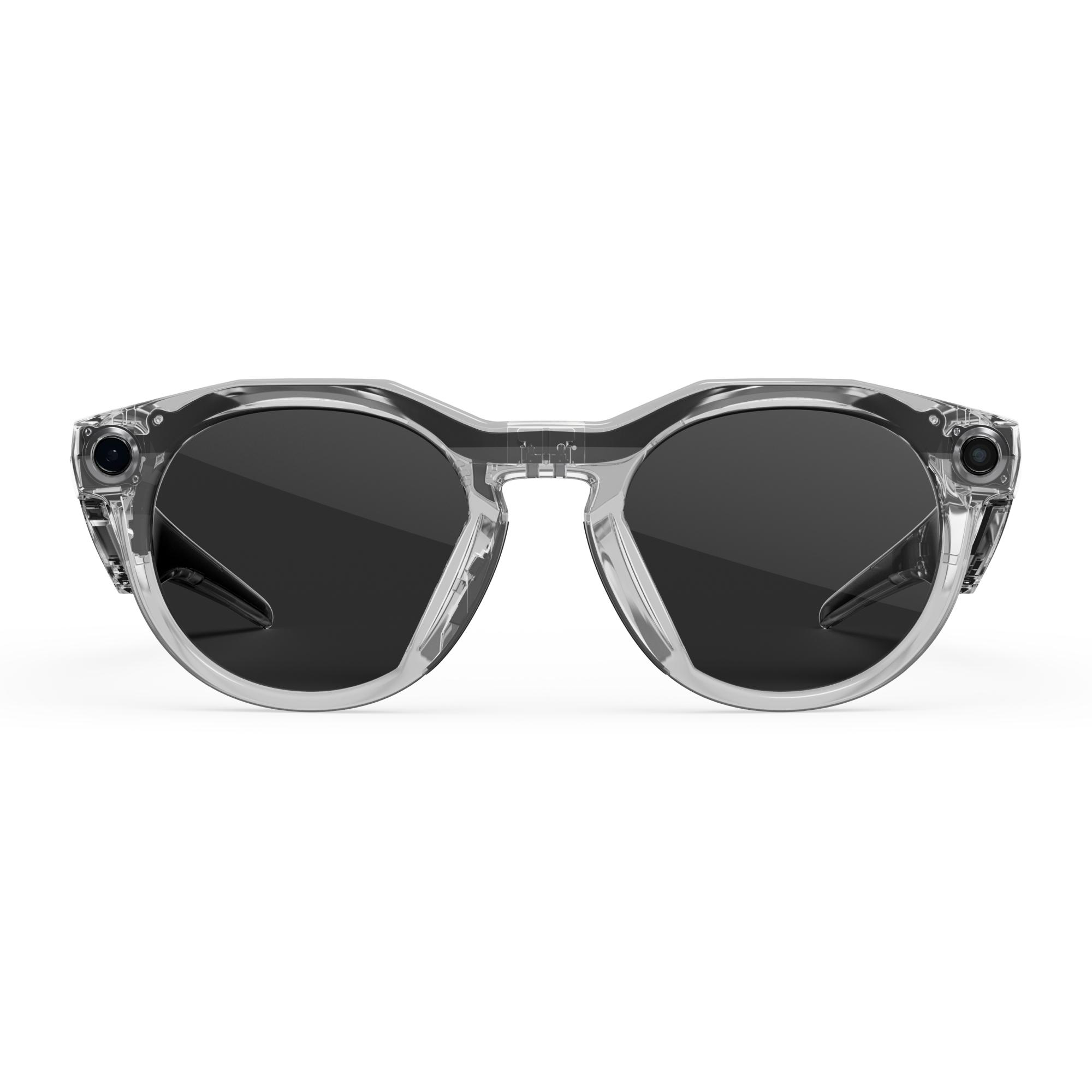 Eine Brille mit durchsichtigem Rahmen und dunklen Gläsern, die sich vor weißem Hintergrund zentriert. Sie hat eine runde Form und Kameras an den Seiten.