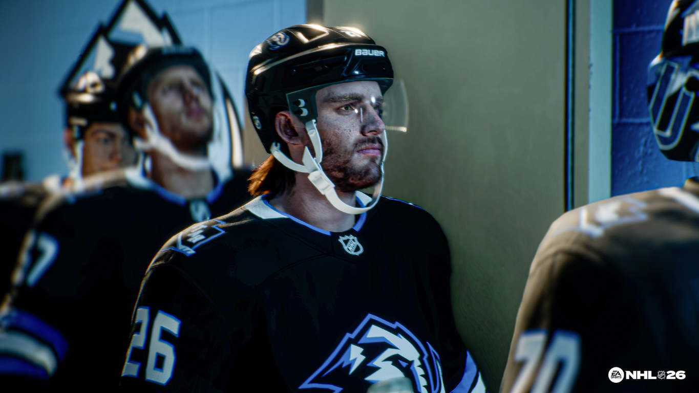 Hokeiści w szatni. Zawodnik w czarnej koszulce z numerem 26 wydaje się skupiony. Inni gracze w tle.