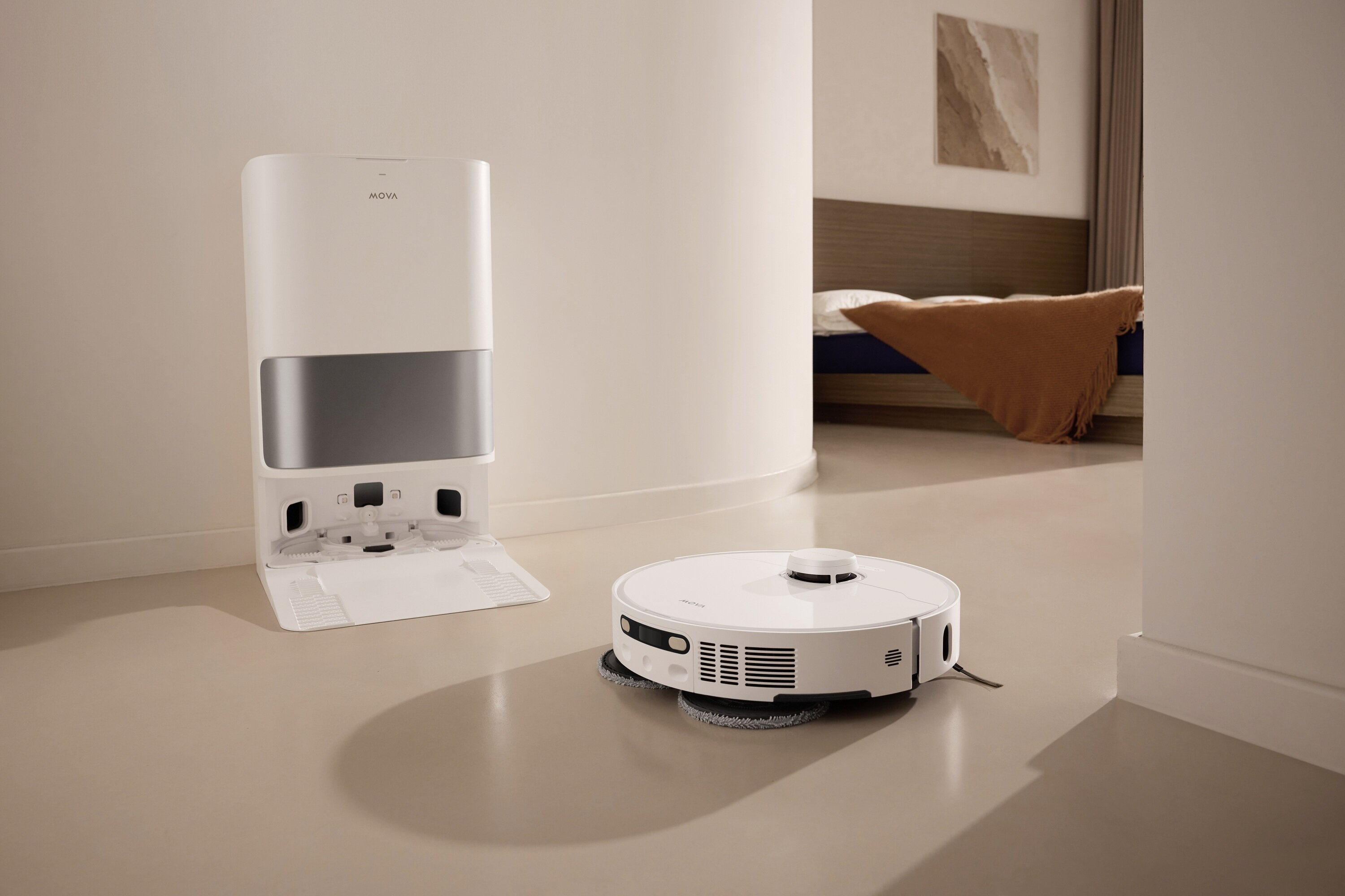 Biały odkurzacz robot i stacja ładująca w pokoju z łóżkiem w tle.
