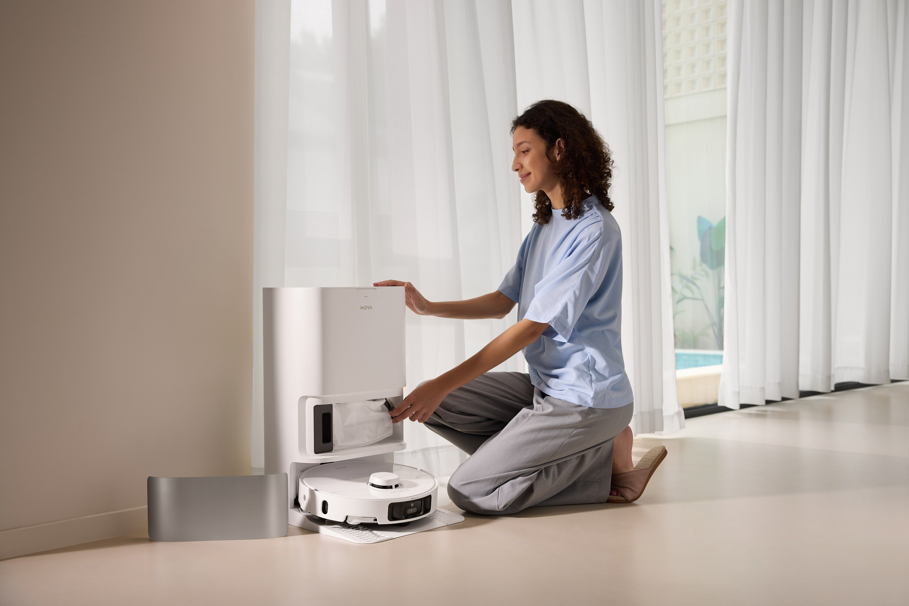 Kobieta klęczy, obsługując robota sprzątającego i jego stację ładującą. Jasnoniebieska koszula, szare spodnie, białe zasłony i jasna podłoga.