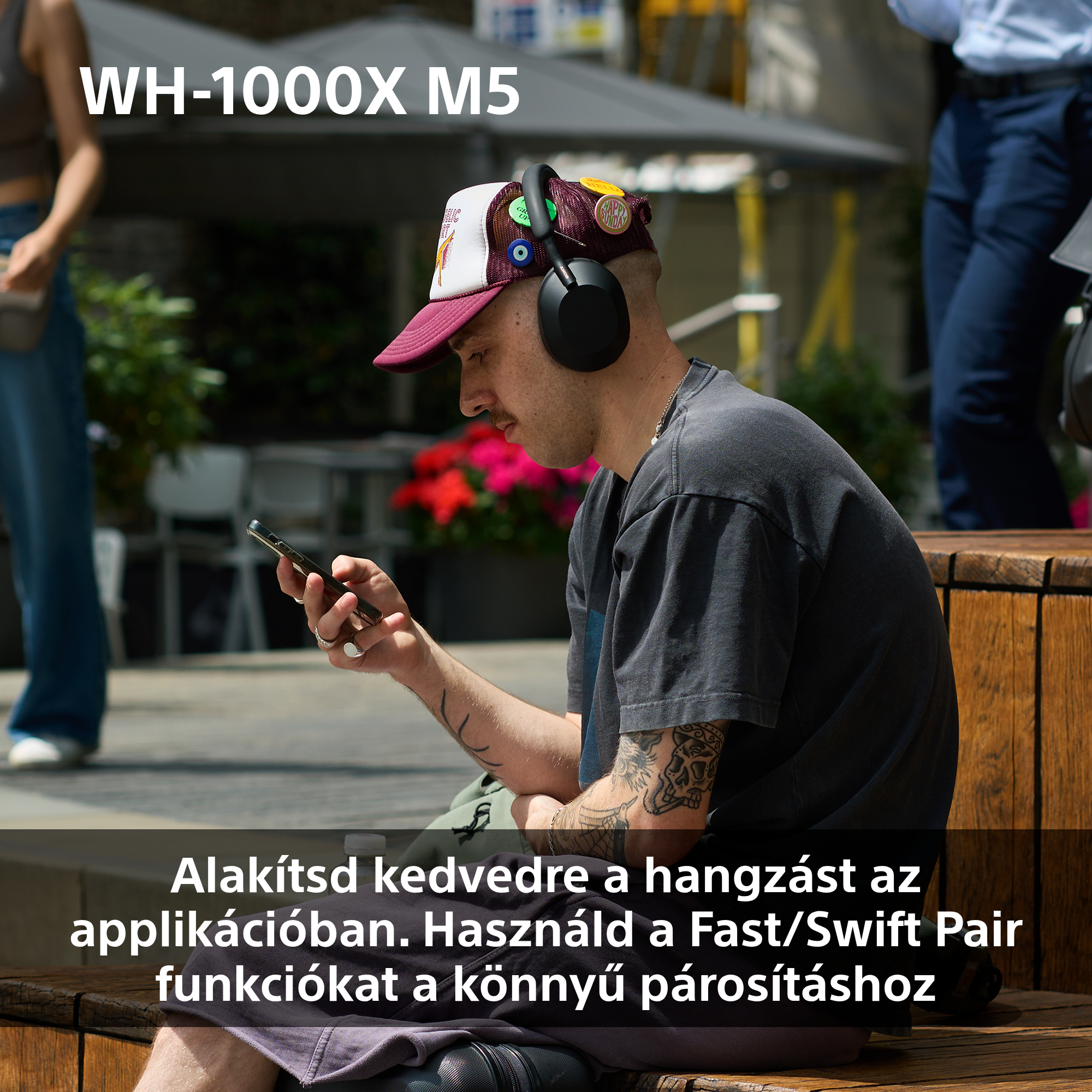 Egy férfi fejhallgatóval és sapkával, telefont használ. A 'WH-1000X M5' terméknév látható. A kép kint készült.