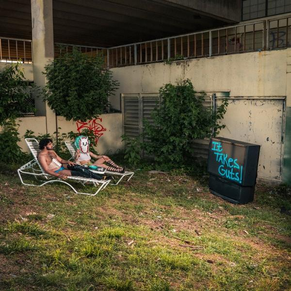 Zwei Personen entspannen sich in Stühlen im Freien. Einer trägt eine Maske. Ein Fernseher zeigt: „It takes guts!“. Graffiti und Grün umgeben sie.