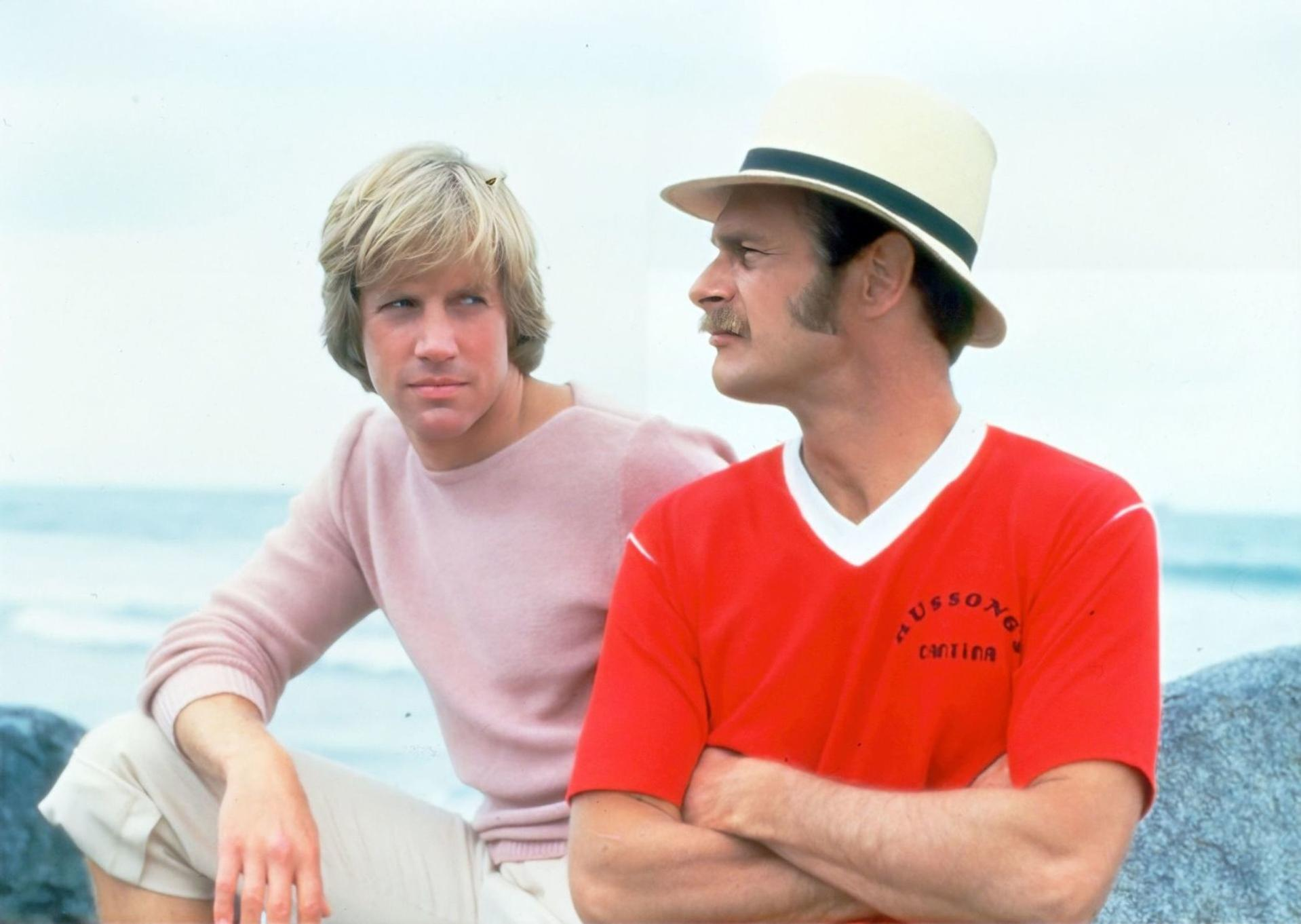 Zwei Männer sitzen draußen. Einer trägt ein rotes Hemd und einen Hut, der andere einen rosa Pullover. Im Hintergrund ist das Meer.