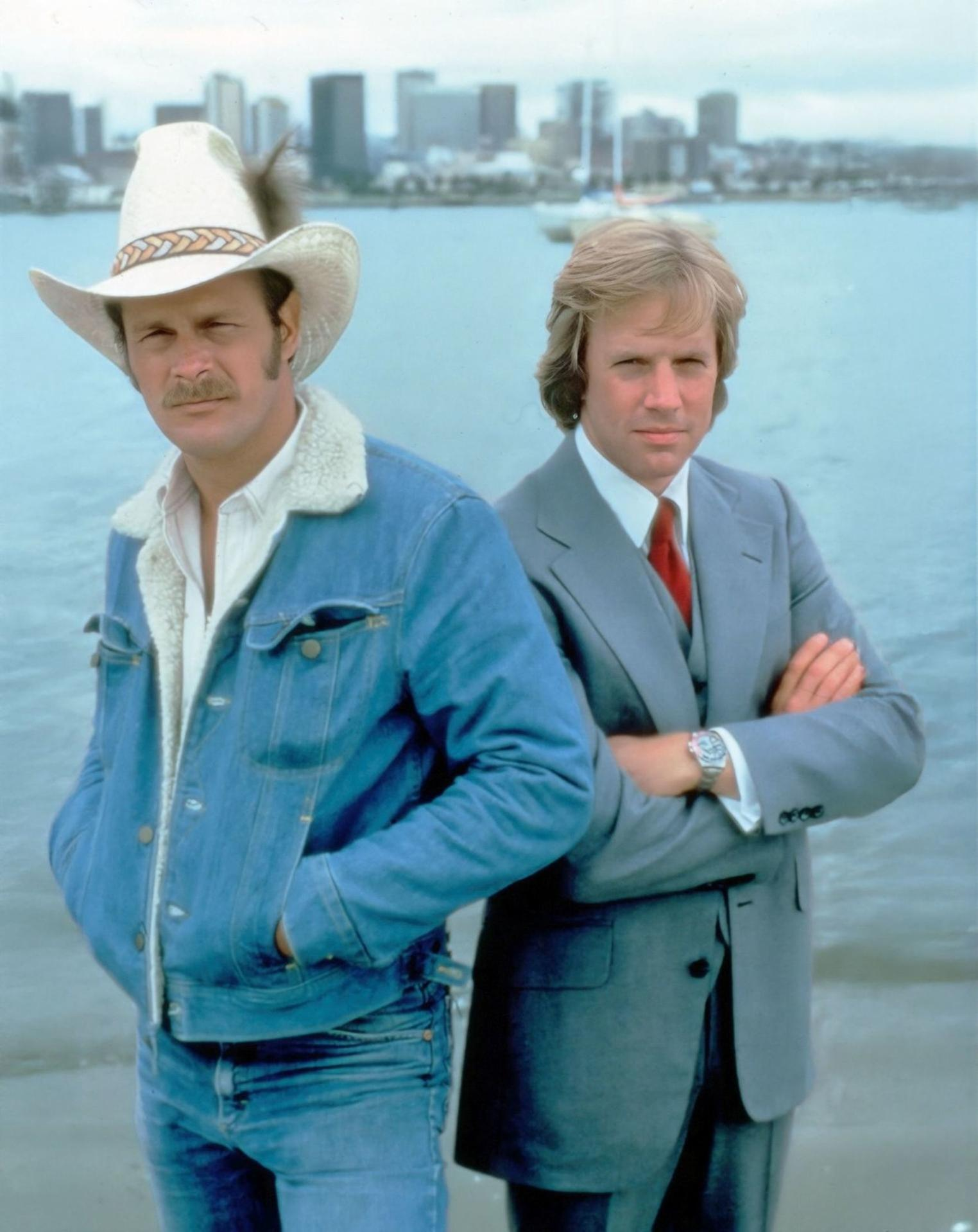 Zwei Männer stehen am Wasser. Einer in Jeansjacke und Cowboyhut, der andere im Anzug, Arme verschränkt. Im Hintergrund eine Stadt.