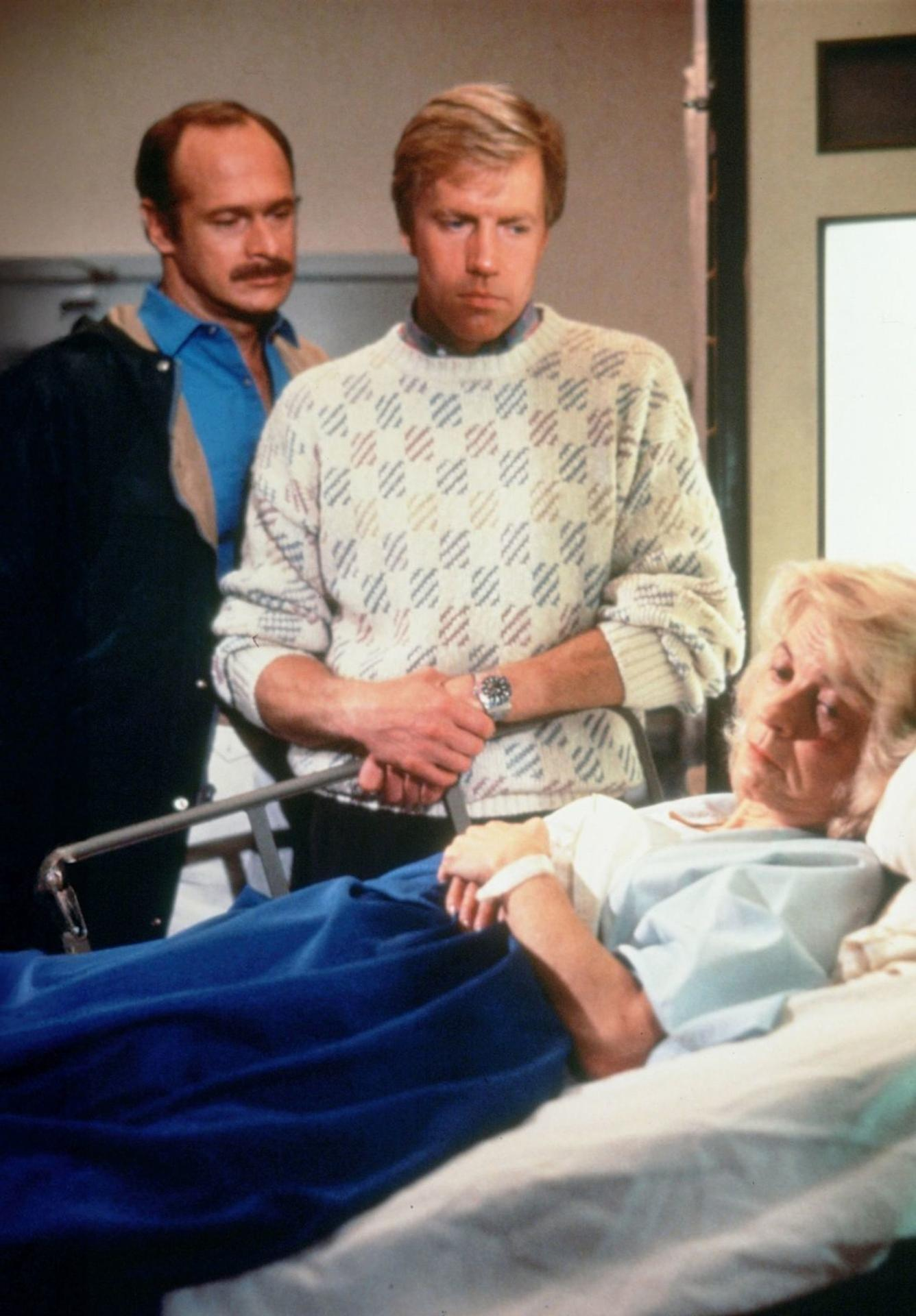 Drei Personen in einem Krankenhauszimmer. Eine ältere Frau liegt im Bett, zwei Männer stehen in ihrer Nähe, einer trägt einen gemusterten Pullover.