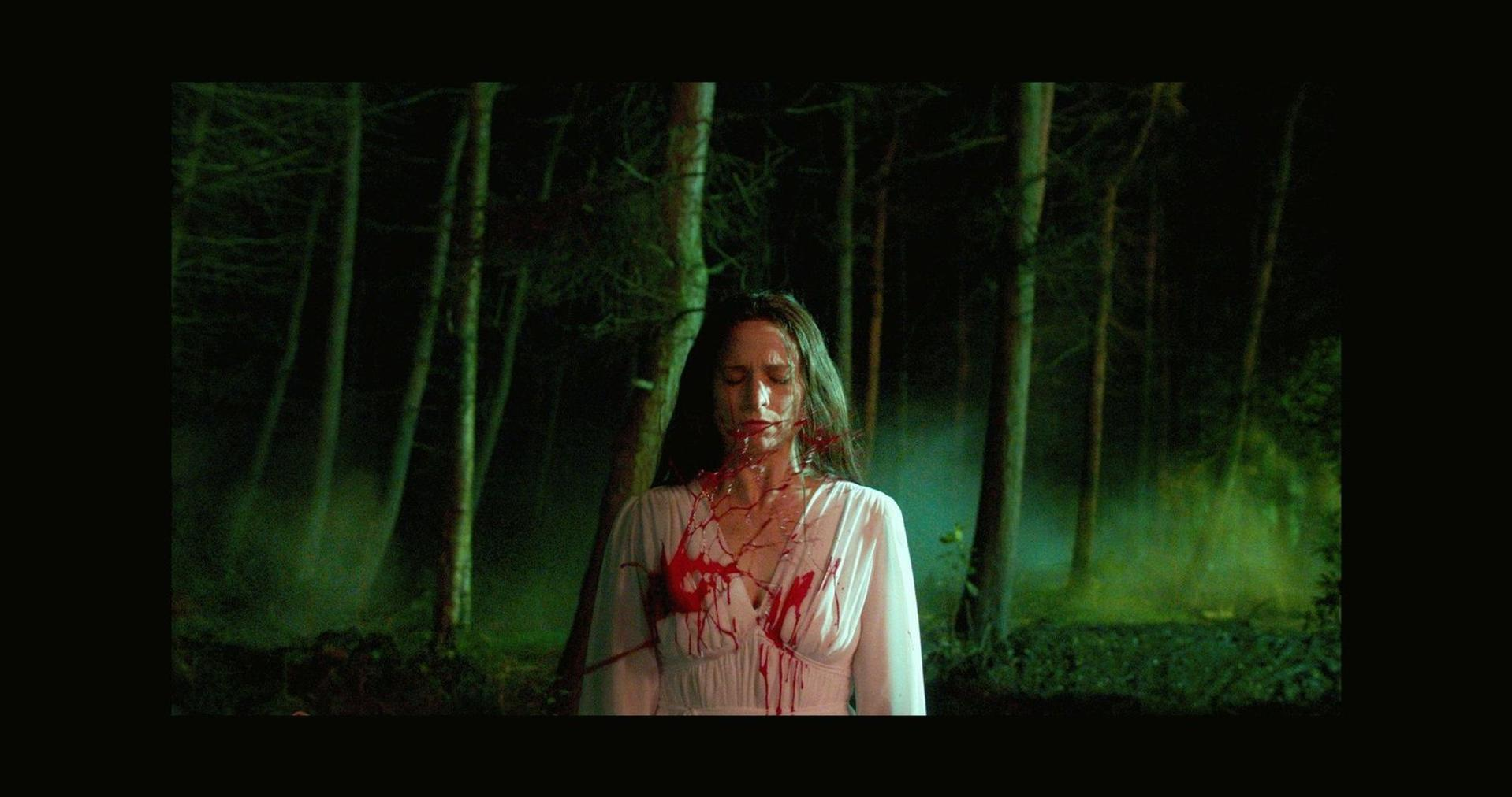 Eine Frau in weißem Kleid steht in einem Wald, bedeckt mit Blut von einer Brustwunde. Bäume bilden den Hintergrund.
