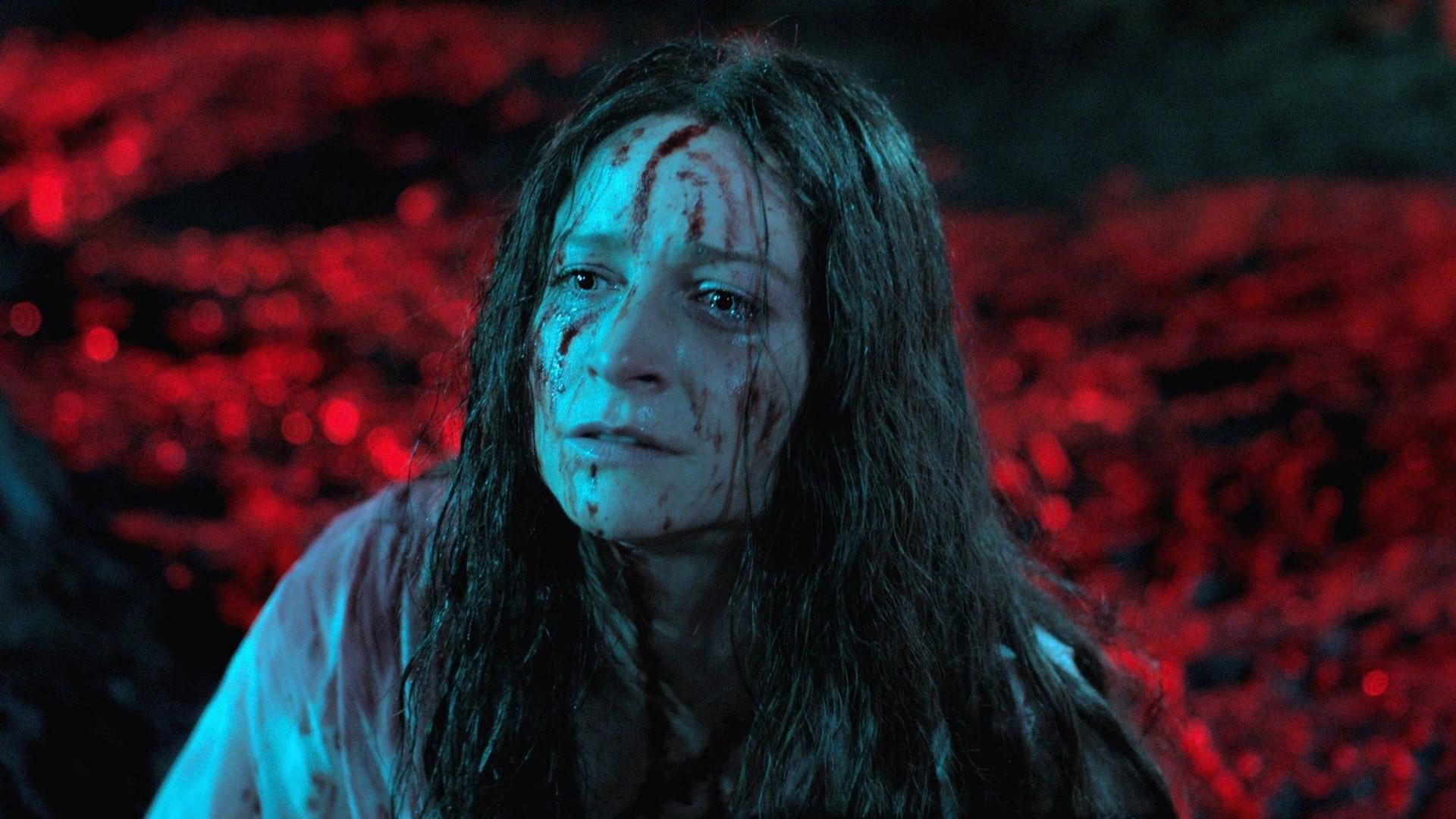 Eine Frau mit Blut im Gesicht weint, dunkle Haare und ein weißes Hemd. Der Hintergrund sind verschwommene rote Lichter.