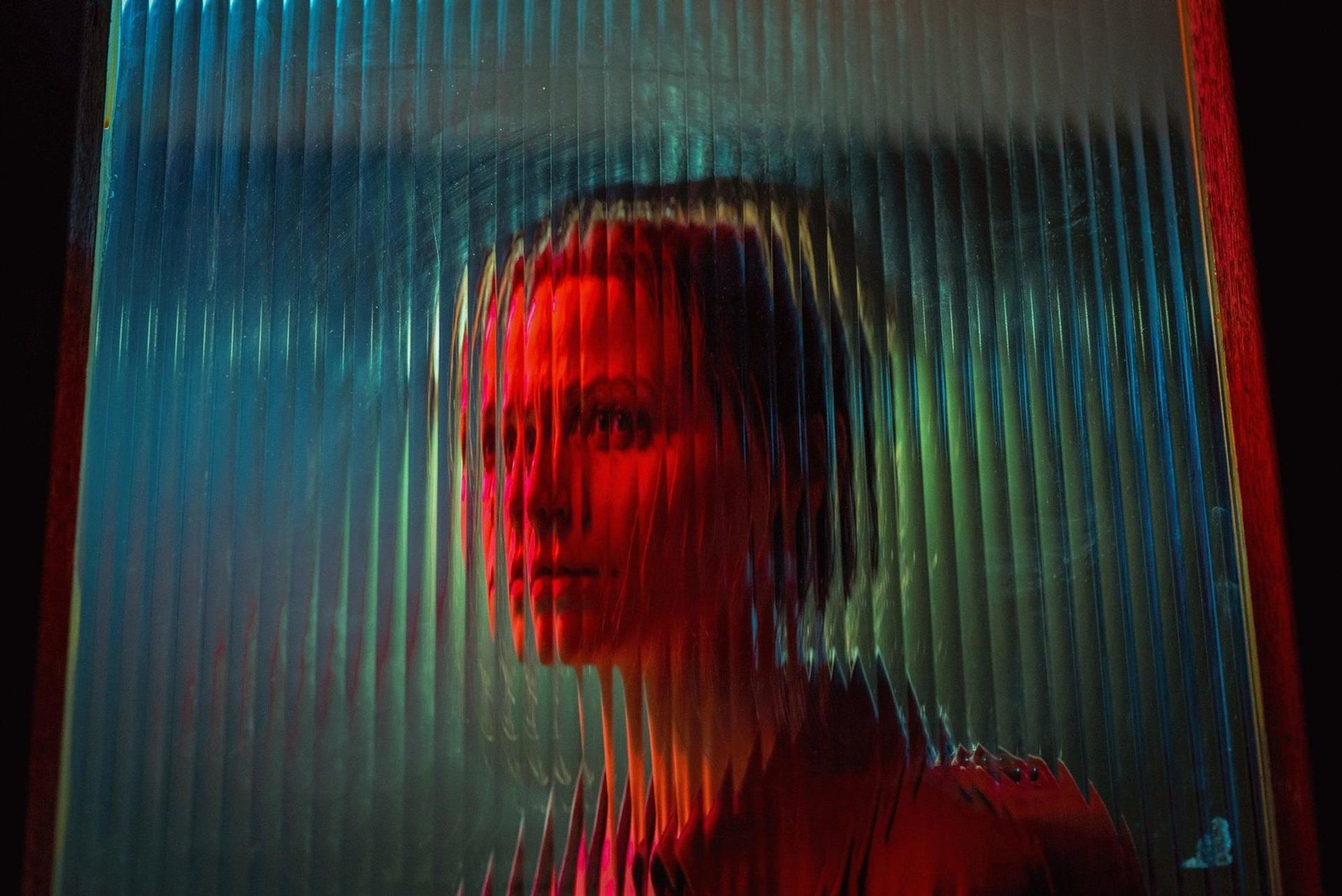 Das Gesicht einer Frau wird durch strukturiertes Glas gesehen. Rotes Licht beleuchtet ihr Gesicht. Der Hintergrund ist blau und grün.