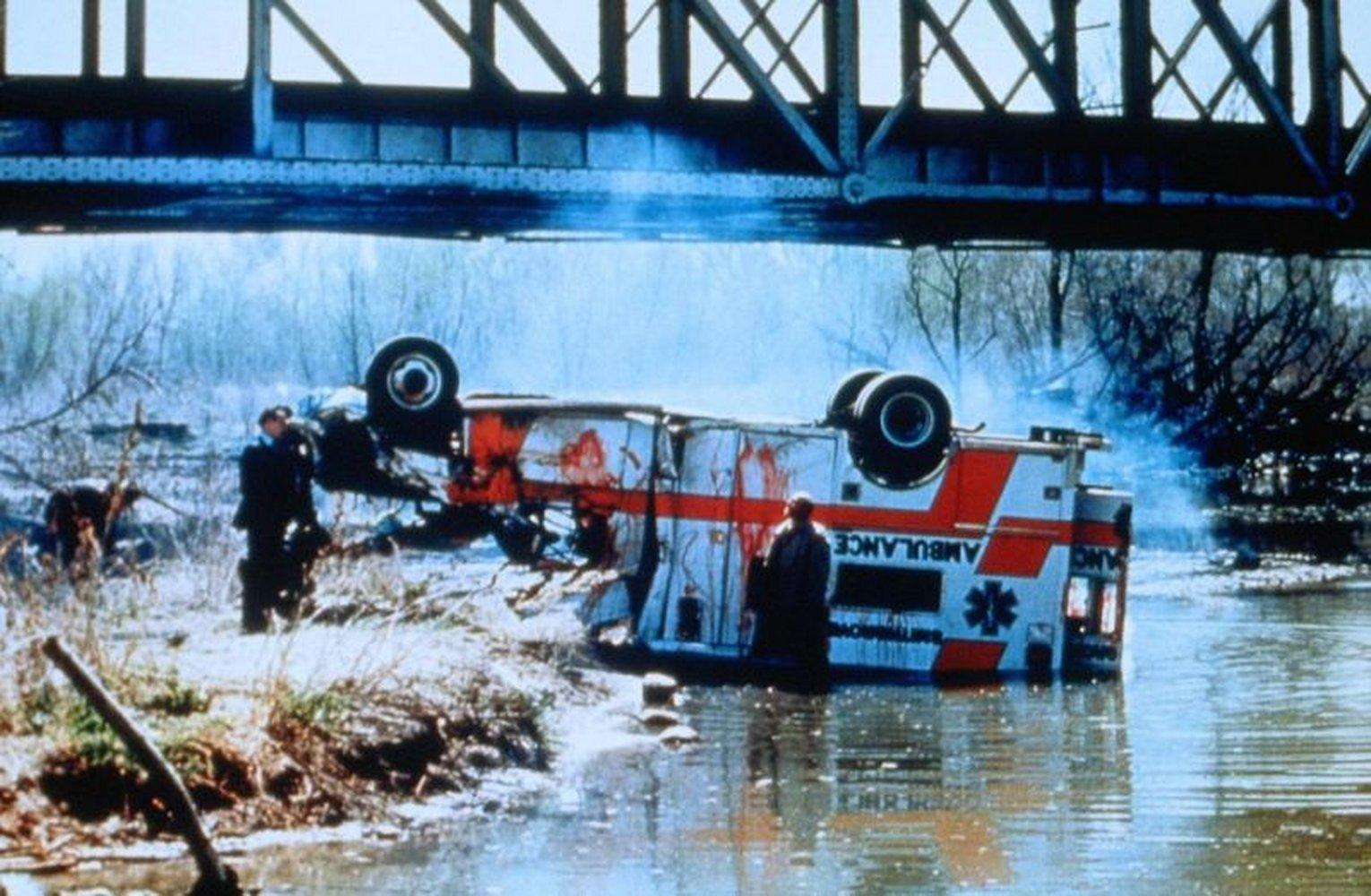 Ein umgestürzter Krankenwagen im Wasser unter einer Brücke. Menschen stehen in der Nähe. Roter und weißer Krankenwagen, schwarze Brücke, blaues Wasser.