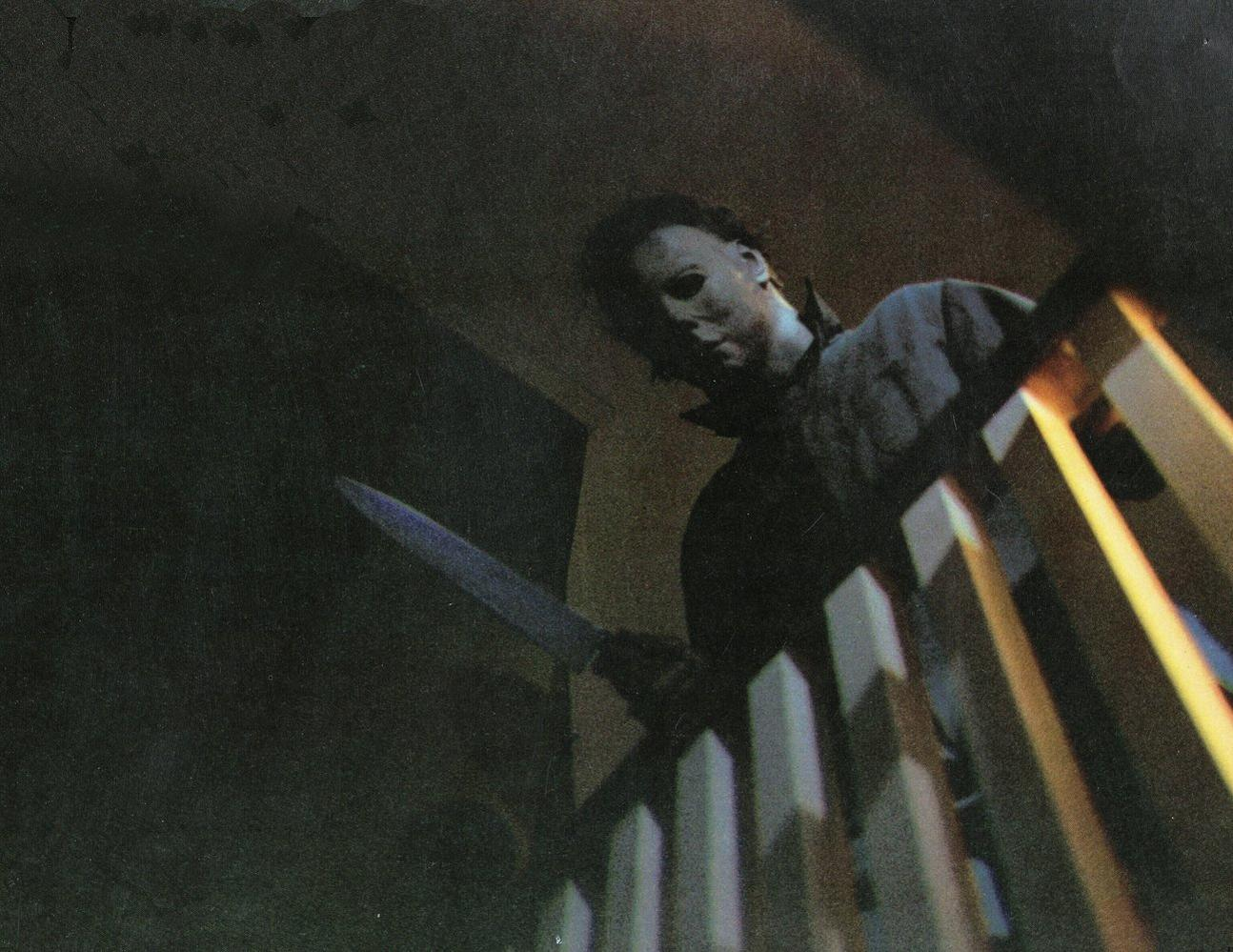 Eine Person mit Maske hält ein Messer auf einer Treppe. Die Beleuchtung ist gedämpft und erzeugt ein Gefühl von Spannung und Vorahnung.