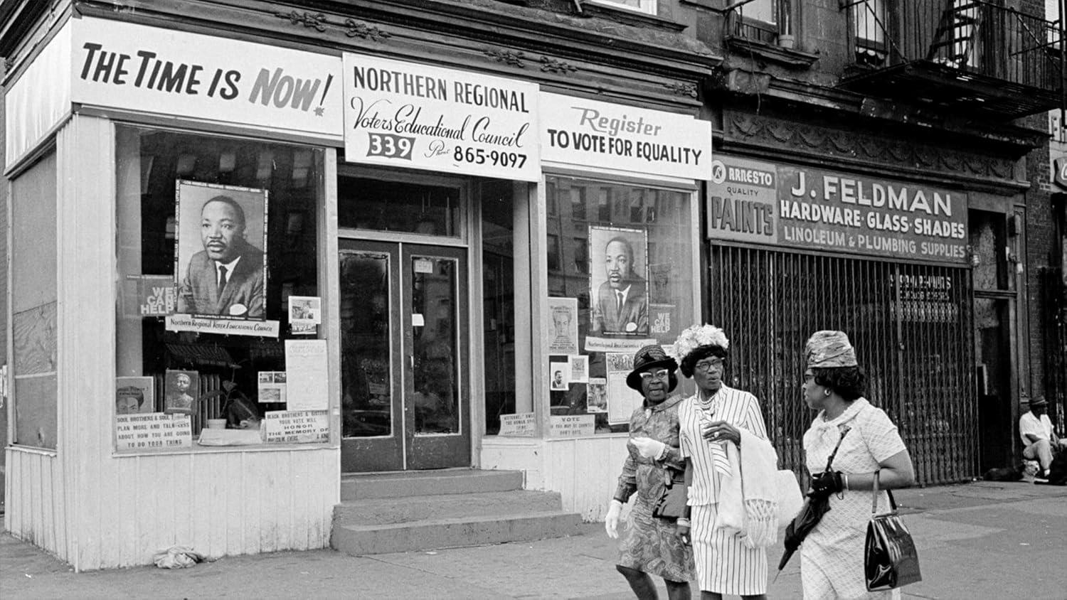 Schwarz-Weiß-Foto eines Ladenschilds mit der Aufschrift „THE TIME IS NOW“. Drei Frauen gehen vorbei. Plakate von Martin Luther King Jr. sind sichtbar.
