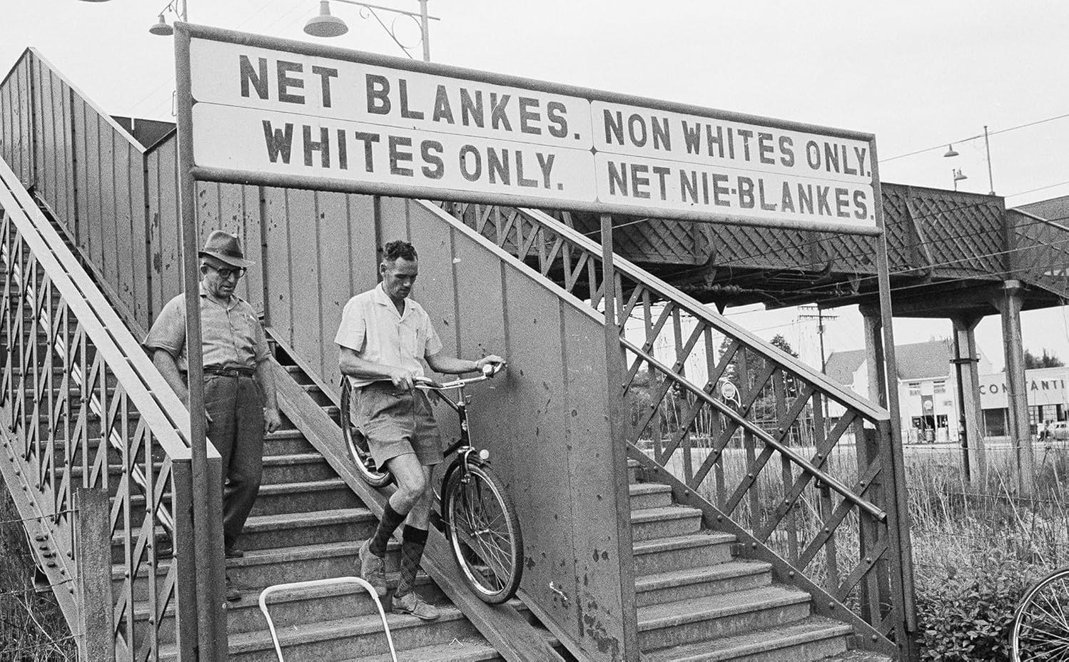 Männer mit einem Fahrrad gehen eine Treppe hinunter, unter einem Schild mit der Aufschrift 'Nur für Weiße'. Die Szene scheint sich in einem getrennten öffentlichen Raum zu befinden.