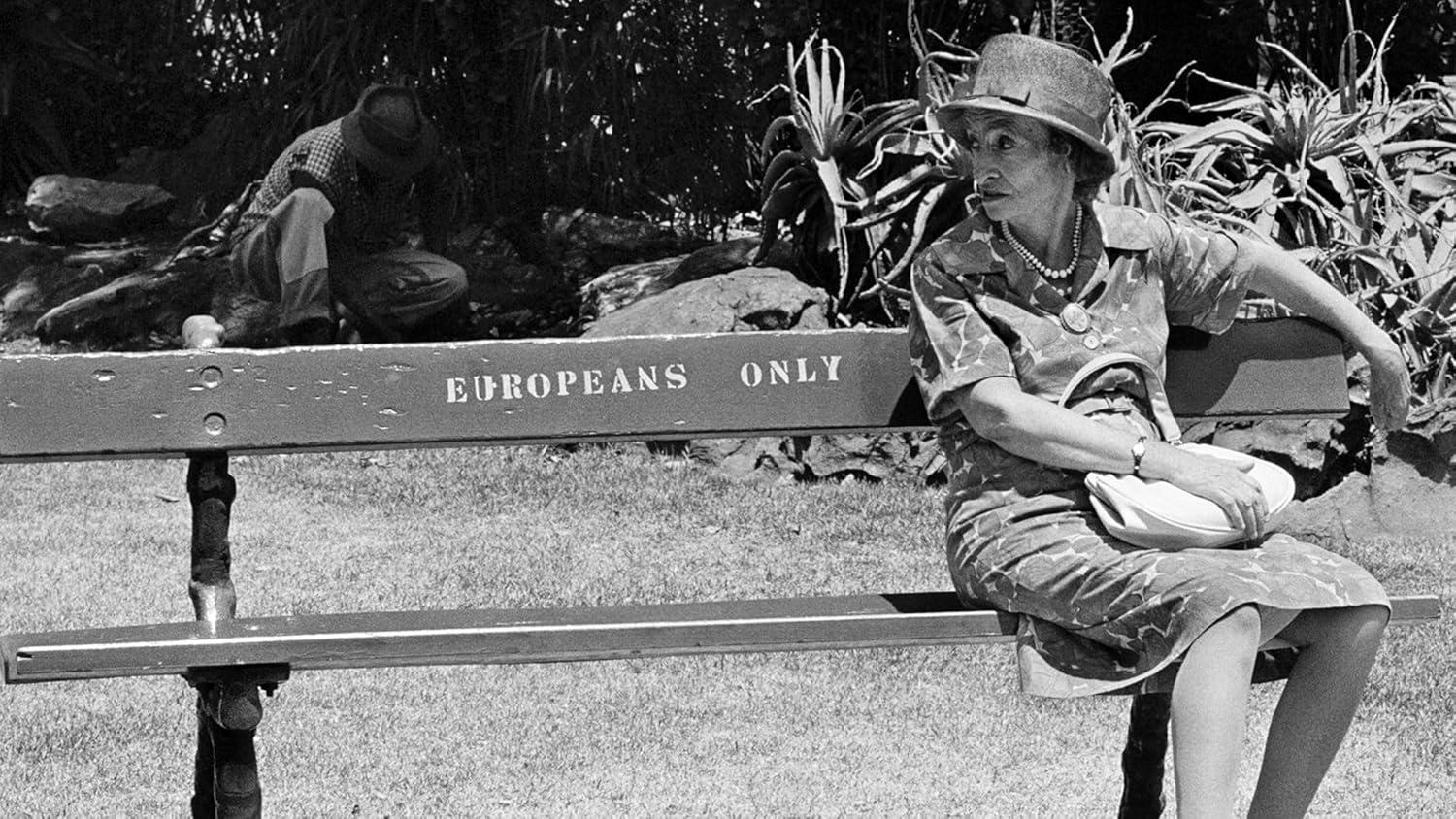 Eine Frau sitzt auf einer Bank mit der Aufschrift 'Europeans Only'. Im Hintergrund ist ein Mann zu sehen. Die Szene ist in Schwarzweiß gehalten.