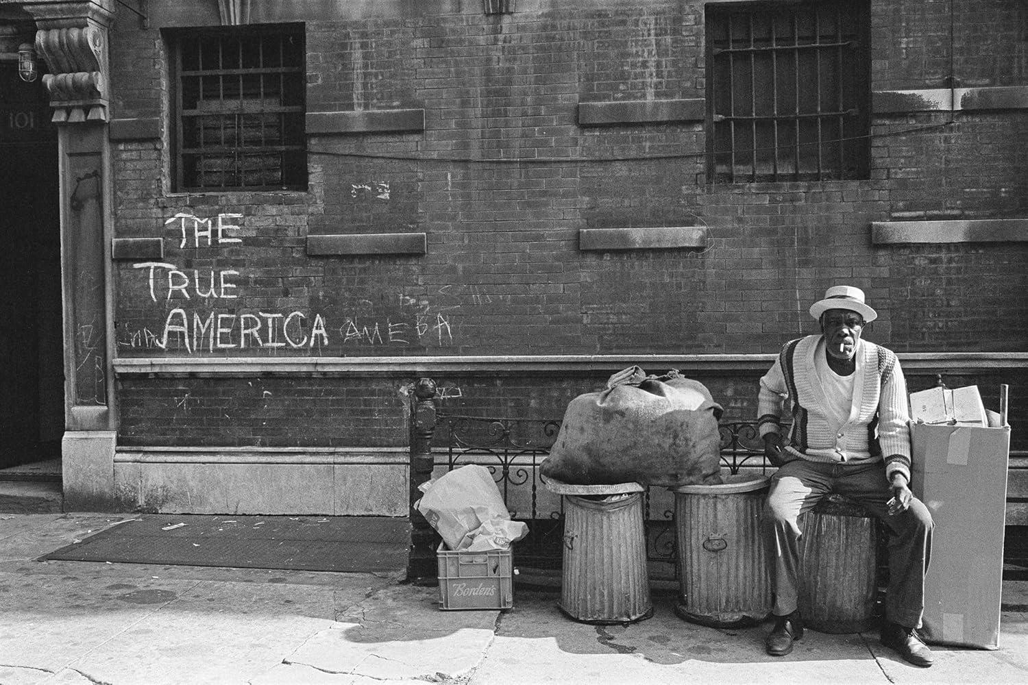 Ein alter Mann sitzt rauchend auf einer Mülltonne. Graffiti auf der Backsteinmauer dahinter lautet: 'The True America'.