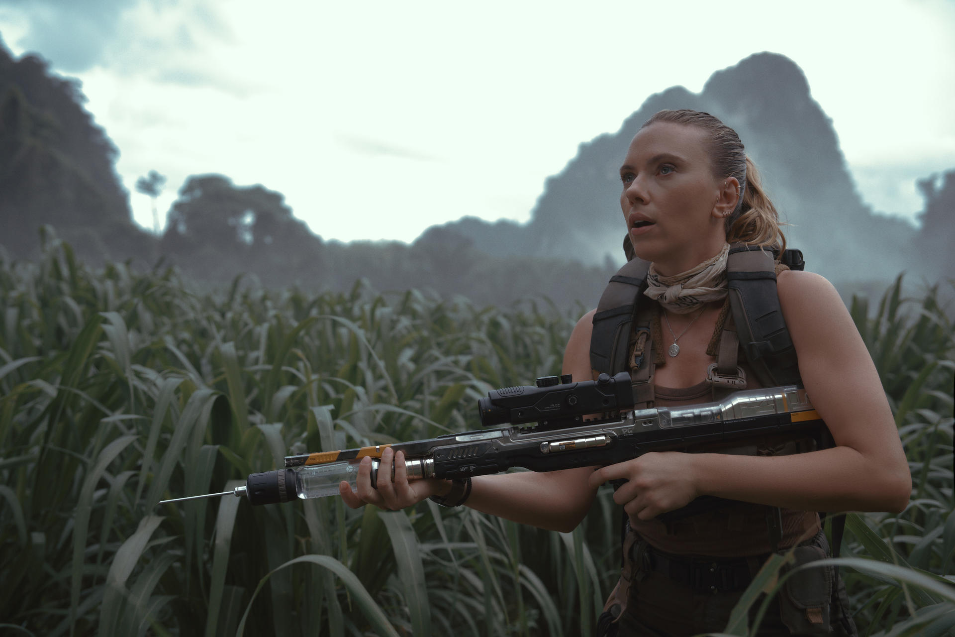 Frau hält eine futuristische Waffe in einem Feld. Berge im Hintergrund. Sie trägt eine Weste und einen Schal und wirkt wachsam.