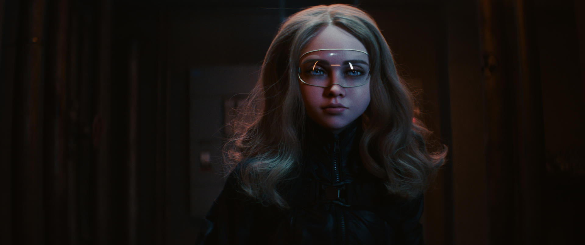 Ein Mädchen mit blonden Haaren und Brille steht in einem dunklen Flur. Sie trägt eine schwarze Jacke. Der Hintergrund ist ein Flur.