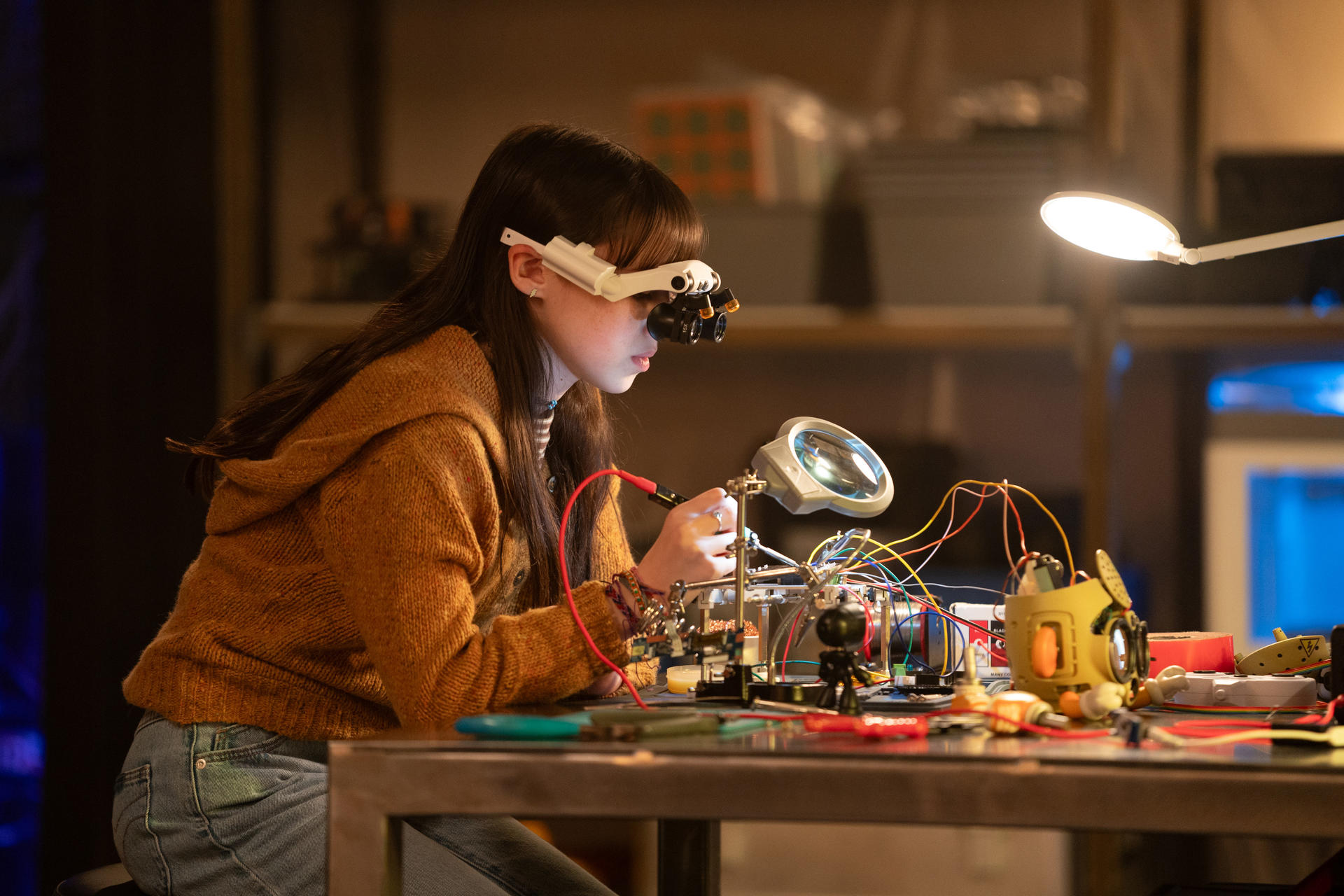 Ein Mädchen mit Schutzbrille lötet Drähte an einem Tisch. Eine Lupe und ein gelber Roboter sind in der Nähe. Im Hintergrund sind Regale.