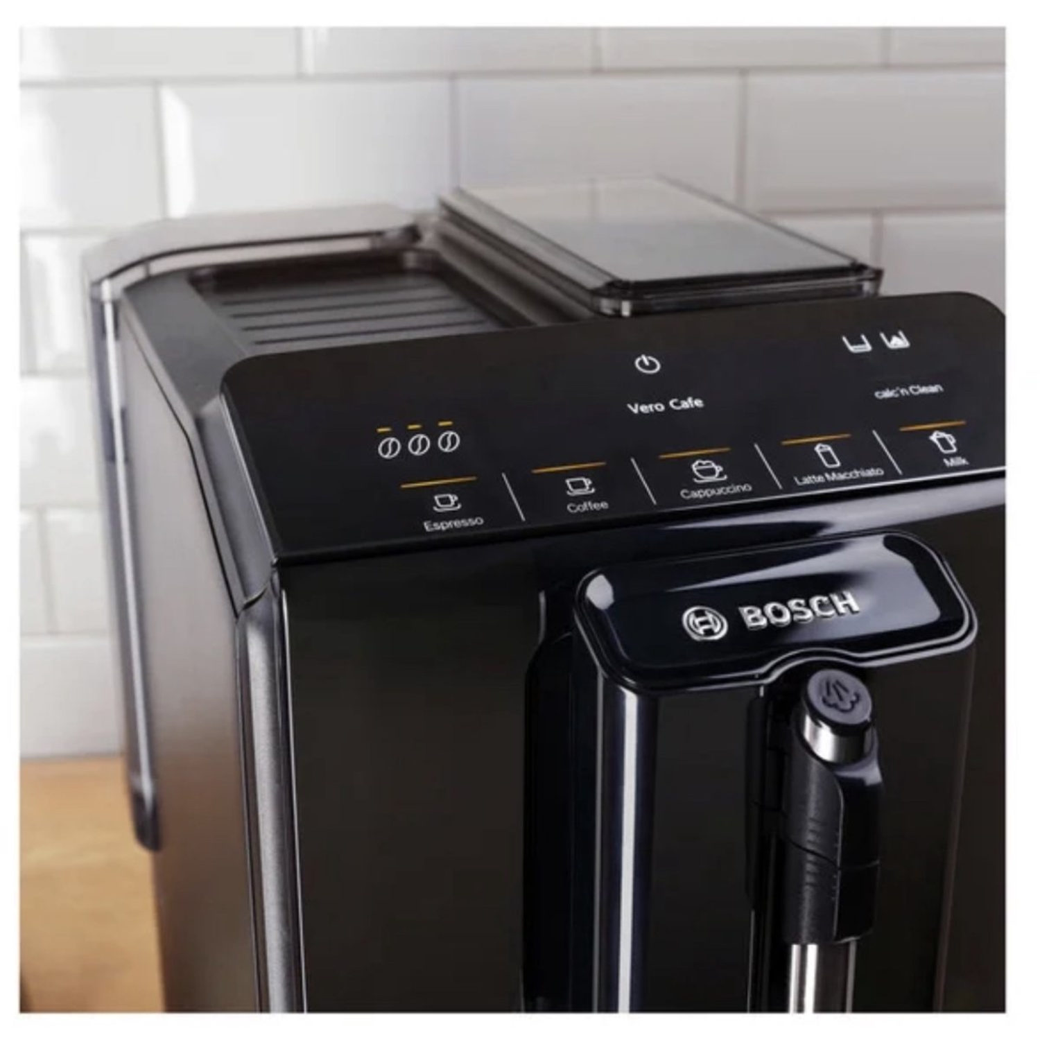 A black Bosch coffee machine with control panel, buttons for coffee options, and a milk frother. The background is a tiled wall.