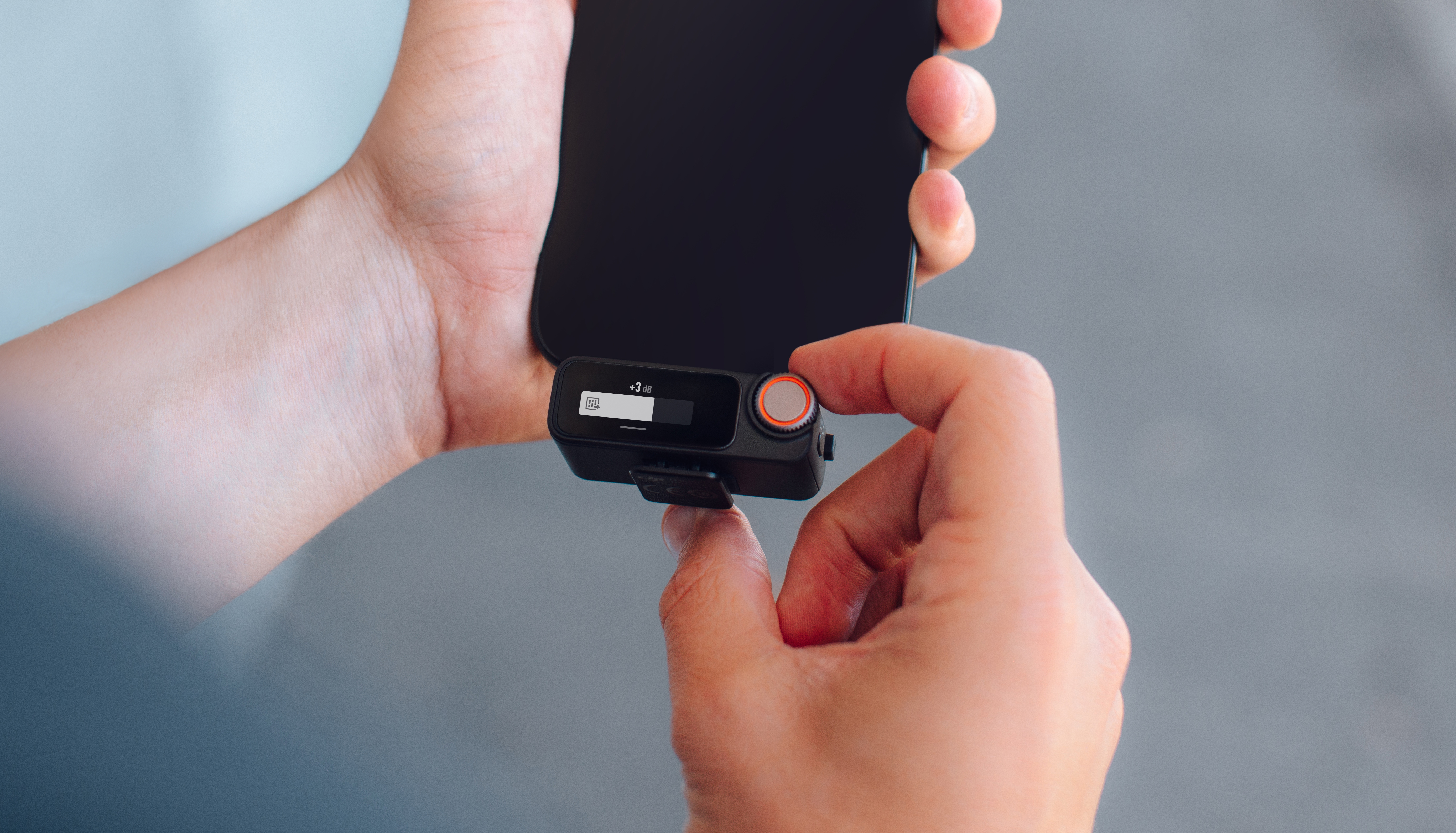 Ręce trzymające czarny smartfon i małe urządzenie audio. Urządzenie ma przycisk głośności i mały ekran.