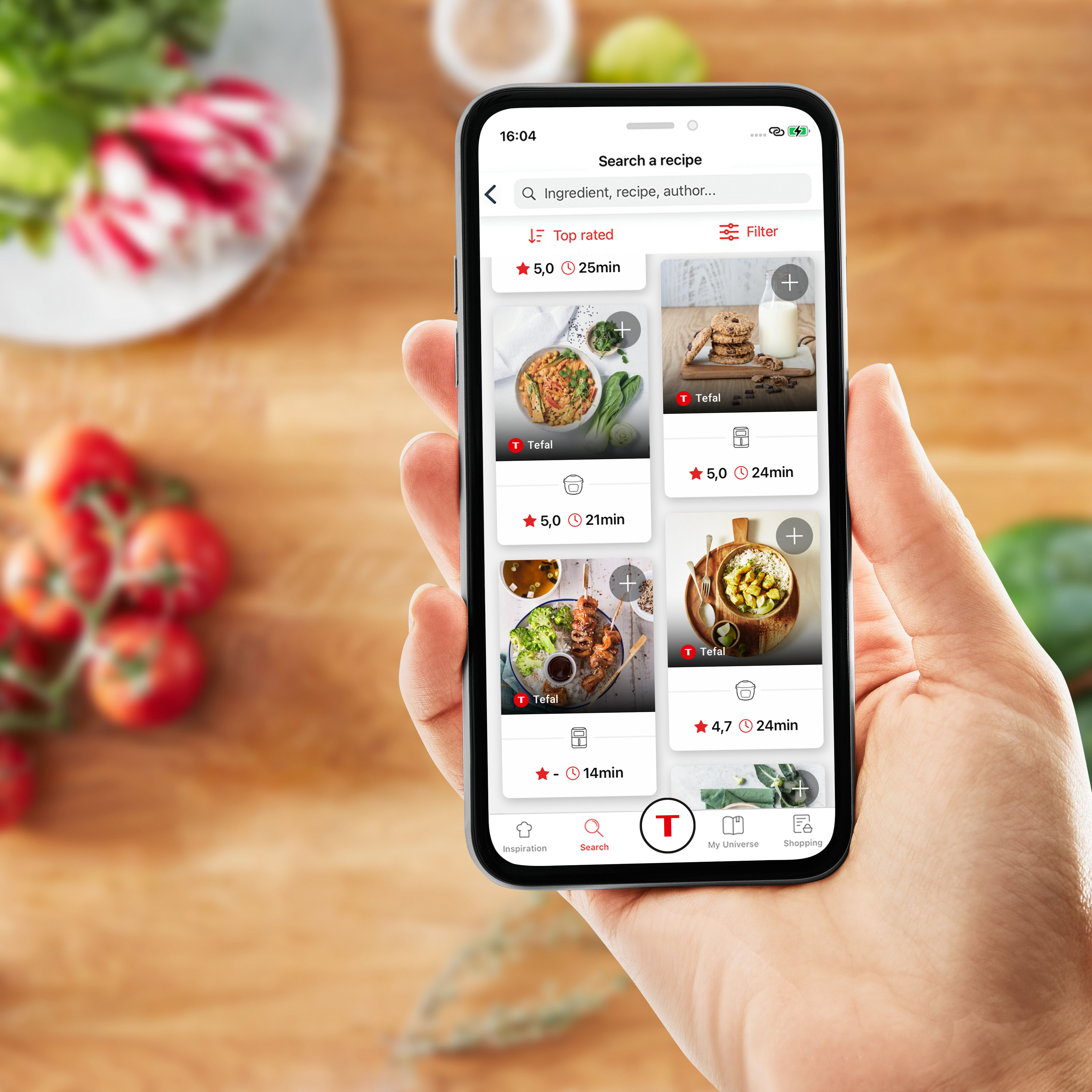 Eine Hand, die ein Telefon mit einer geöffneten Rezept-App hält. Zutaten sind im Hintergrund verschwommen.