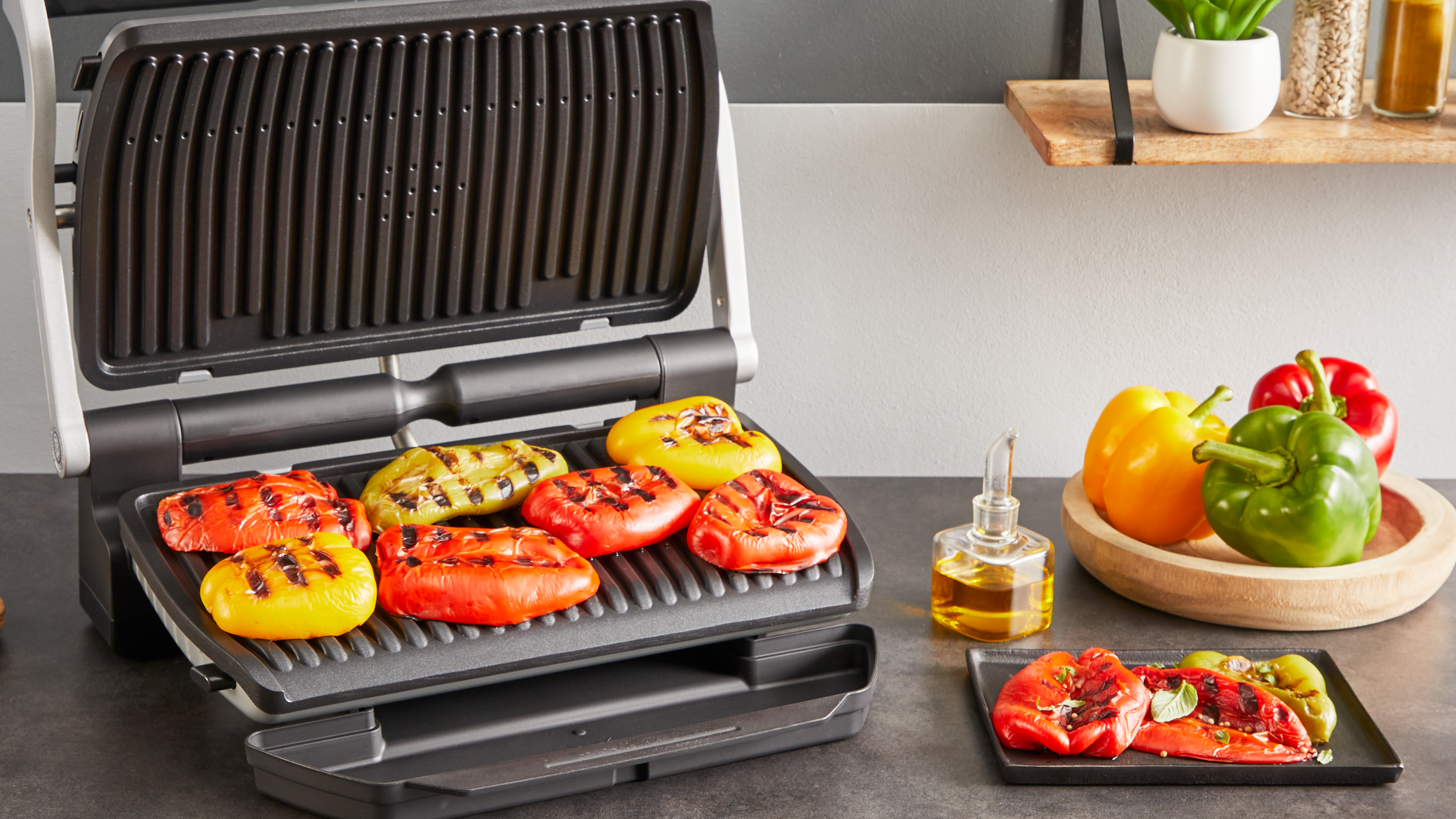 Ein offener Grill mit gegrillten Paprika. Gelbe, rote und grüne Paprika befinden sich auf dem Grill und in einer Holzschale. Öl und Pflanzen sind sichtbar.