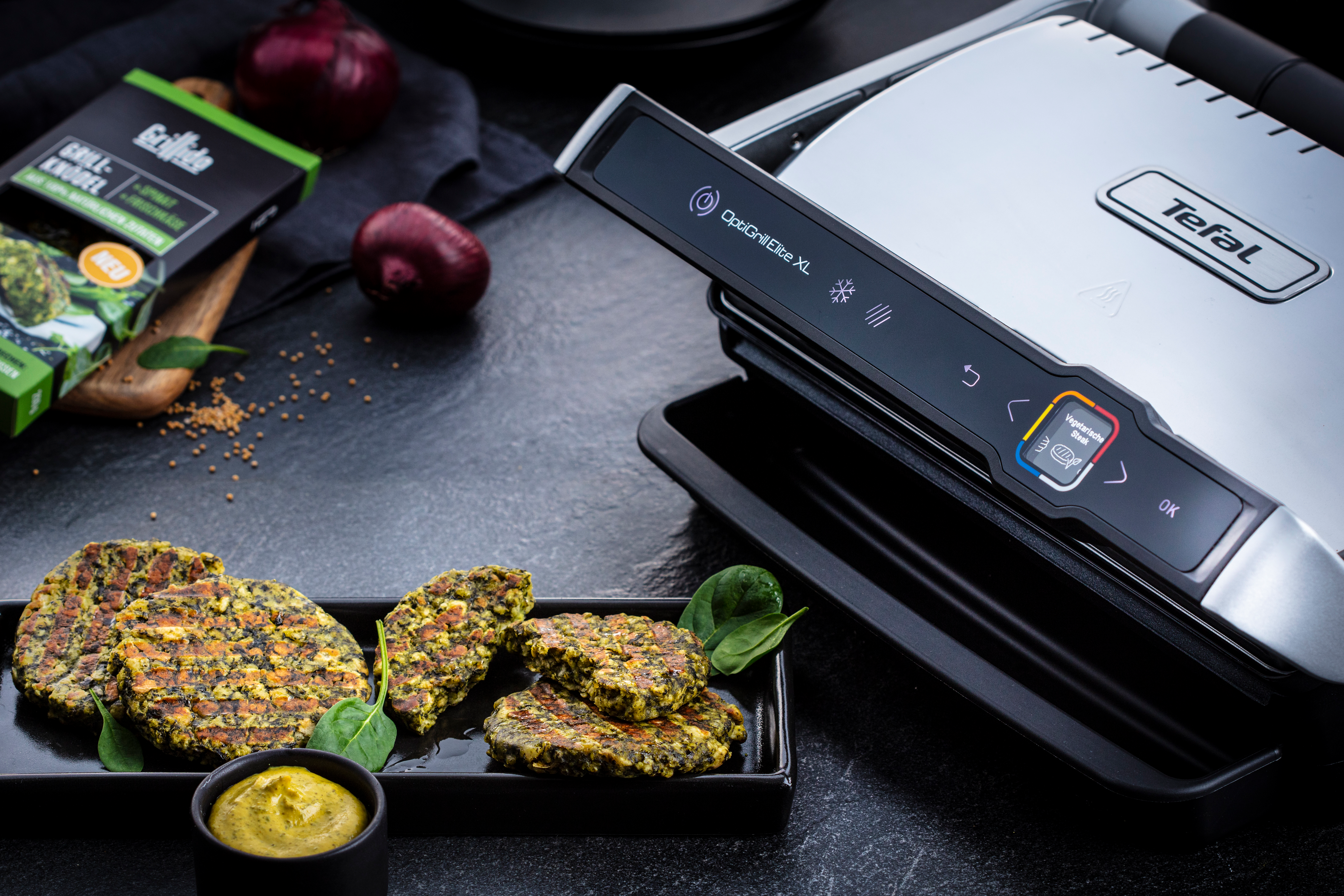 Gegrillte Bratlinge auf einem schwarzen Teller mit Soße, in der Nähe eines Tefal OptiGrill und Lebensmittelverpackungen.