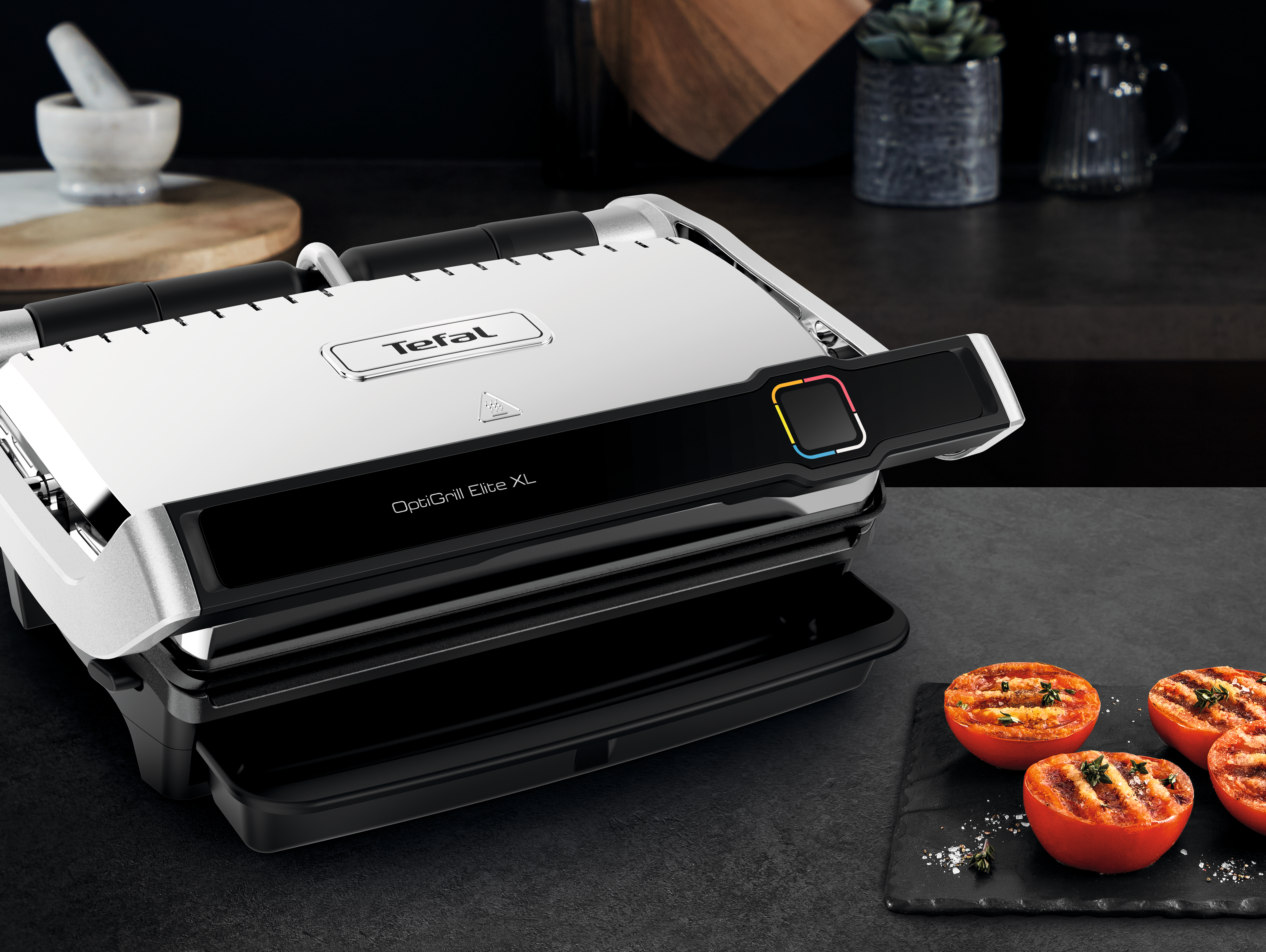 Ein silberner Tefal OptiGrill Elite XL steht auf einer schwarzen Oberfläche. Gegrillte Tomaten auf einem Schieferbrett im Vordergrund.