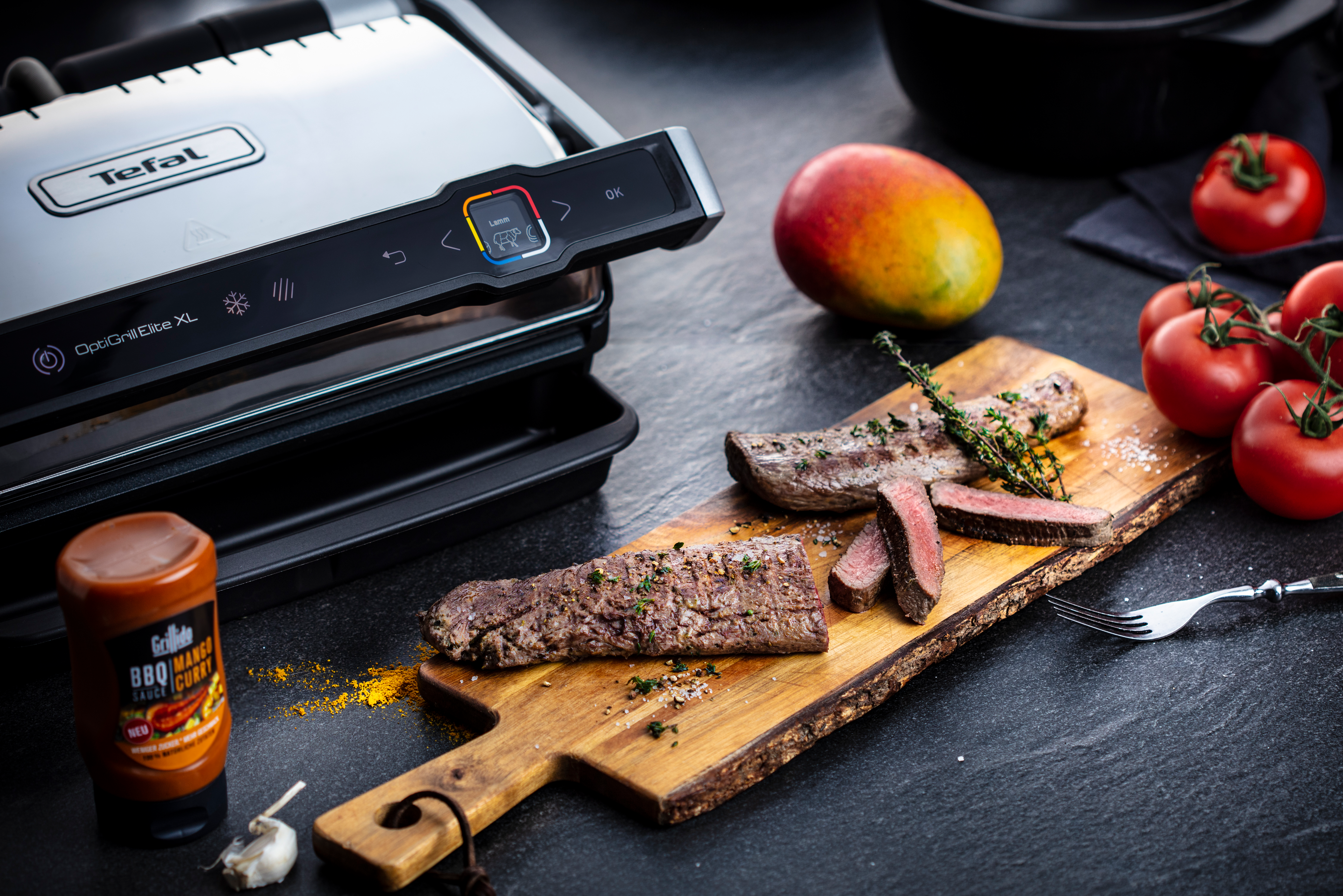 Gegrilltes Steak auf einem Holzbrett mit Tomaten, Mango und einem Tefal OptiGrill Elite XL.