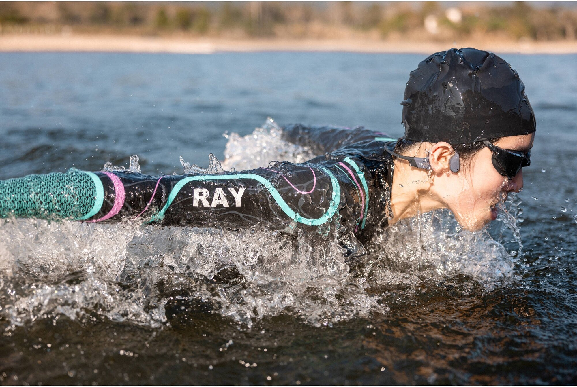 Pływak w wodzie z goglami i czepkiem, logo Ray na kostiumie. Woda pluska wokół. Tło pokazuje wodę i odległą linię brzegową.