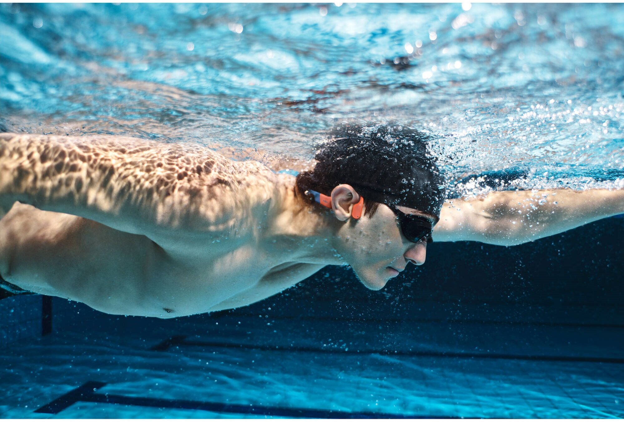 Pływak pod wodą w basenie, w okularach i słuchawkach, wyciągający ręce. Scena jest czysto niebieska.