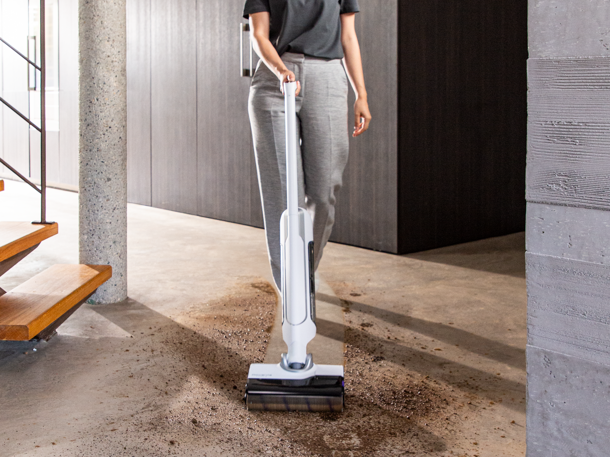 Eine Person saugt einen schmutzigen Boden mit einem weißen Staubsauger. Hintergrund: Treppe, Säulen und dunkle Wand.