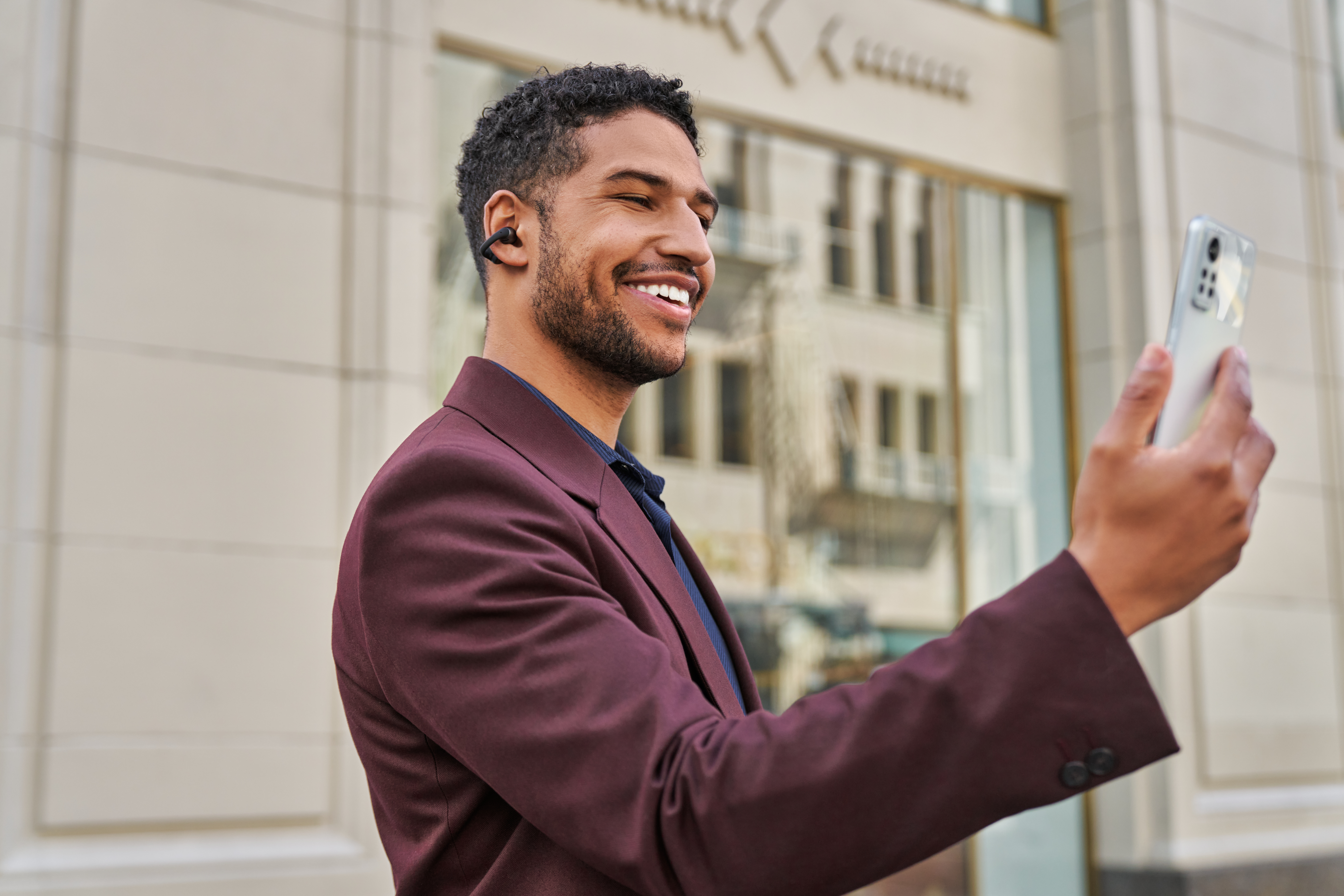 Mężczyzna w bordowym garniturze uśmiecha się, robiąc selfie smartfonem. Nosi bezprzewodową słuchawkę. Budynki w tle.
