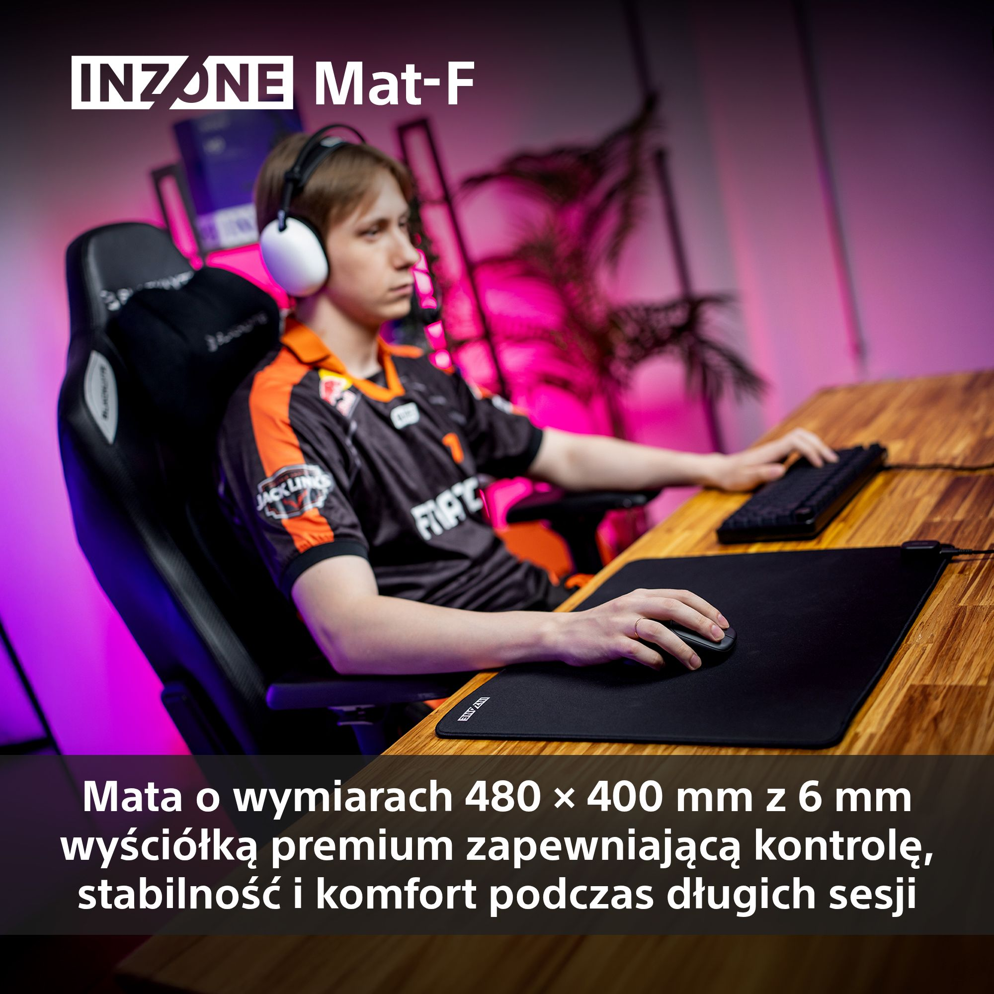 Osoba w słuchawkach siedzi przy biurku z podkładką pod mysz i klawiaturą. Biurko jest drewniane.