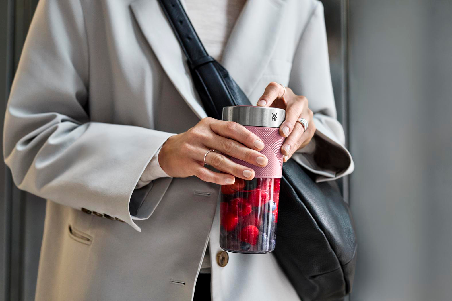 Eine Person hält einen rosa-silbernen Smoothie-Becher mit Beeren, trägt einen grauen Blazer und eine schwarze Tasche.