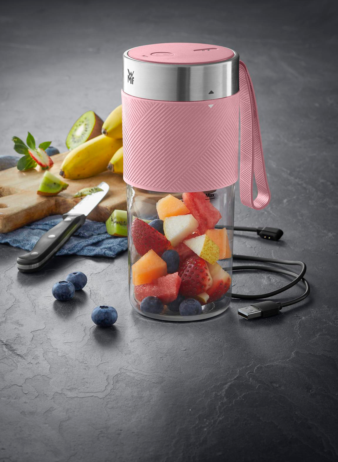 Rosa Mixer gefüllt mit Obst. Erdbeeren, Melone, Wassermelone und Blaubeeren. Bananen und Kiwi auf einem Holzbrett.