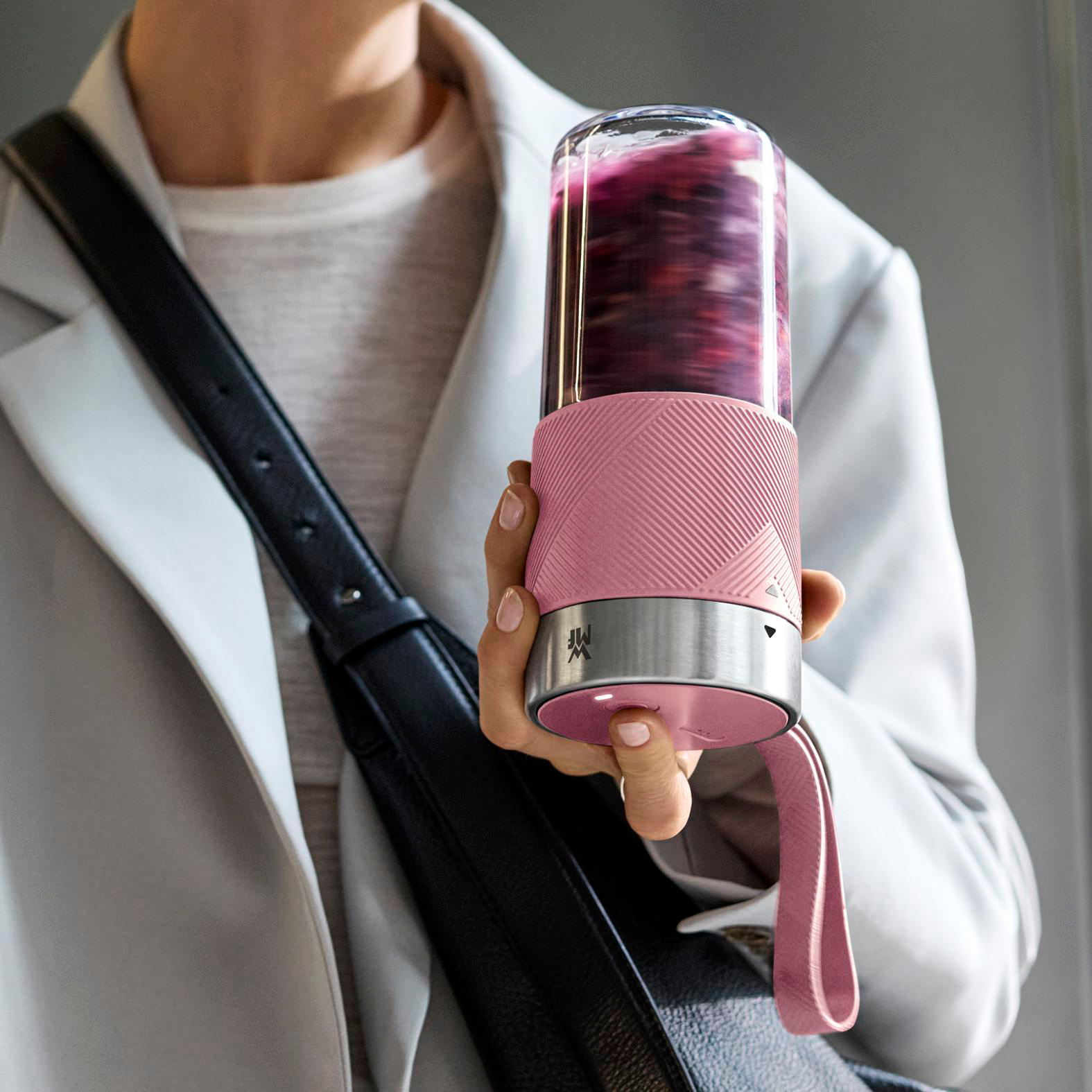 Eine Person hält einen rosa Mixer mit einer violetten Flüssigkeit. Sie trägt einen weißen Mantel und eine schwarze Tasche.