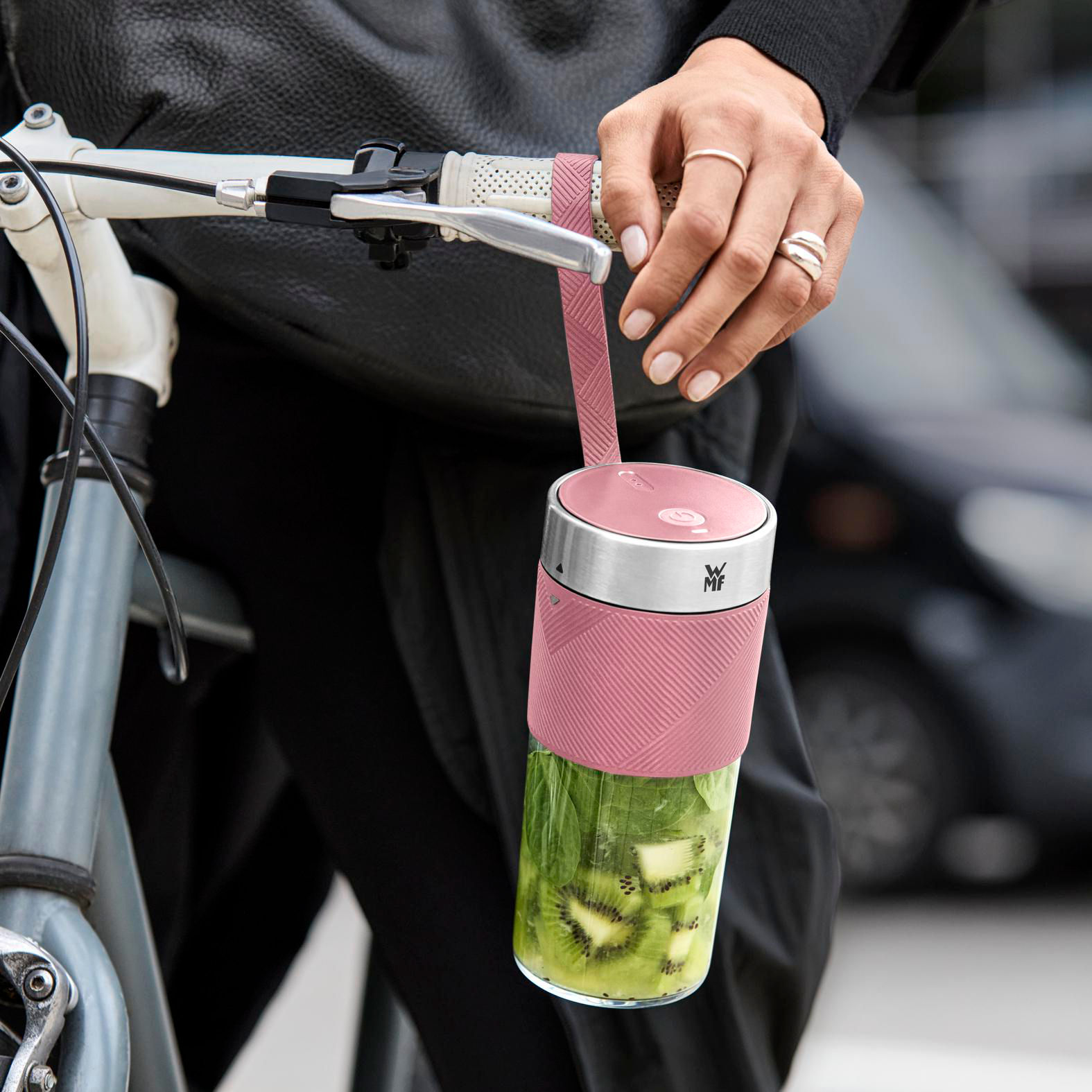 Person hält einen pinkfarbenen Mixer mit Kiwimix, während sie sich auf einen Fahrradlenker stützt. Hintergrund zeigt Straße und Autos.