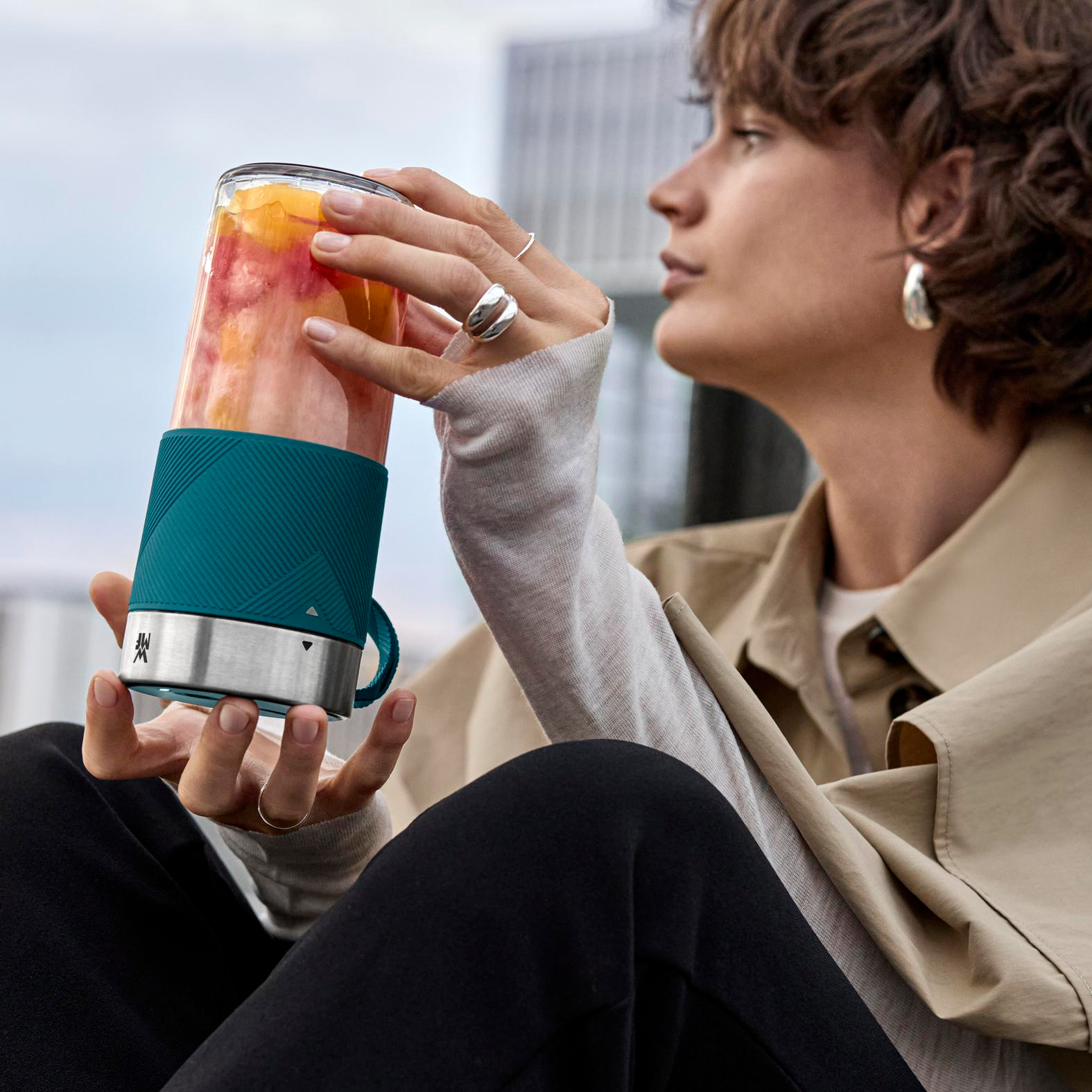 Eine Frau hält einen Smoothie-Maker. Der Maker ist türkis und silber. Der Smoothie im Inneren ist rosa und gelb.