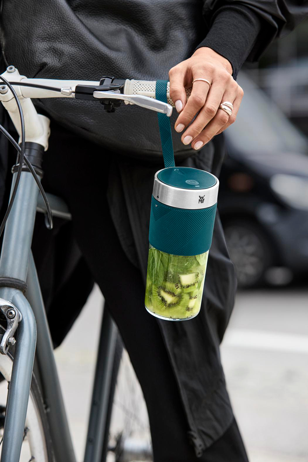 Eine Person hält einen Mixer mit Kiwi und Spinat auf einem Fahrrad. Der Mixer ist türkis und silber. Die Person trägt Ringe und eine schwarze Jacke.