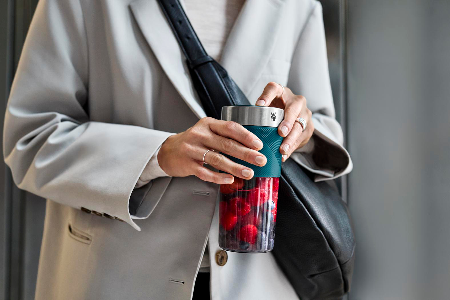 Eine Person hält einen Becher mit Beeren. Sie trägt einen grauen Blazer und eine schwarze Tasche. Der Becher hat einen tealfarbenen Griff.
