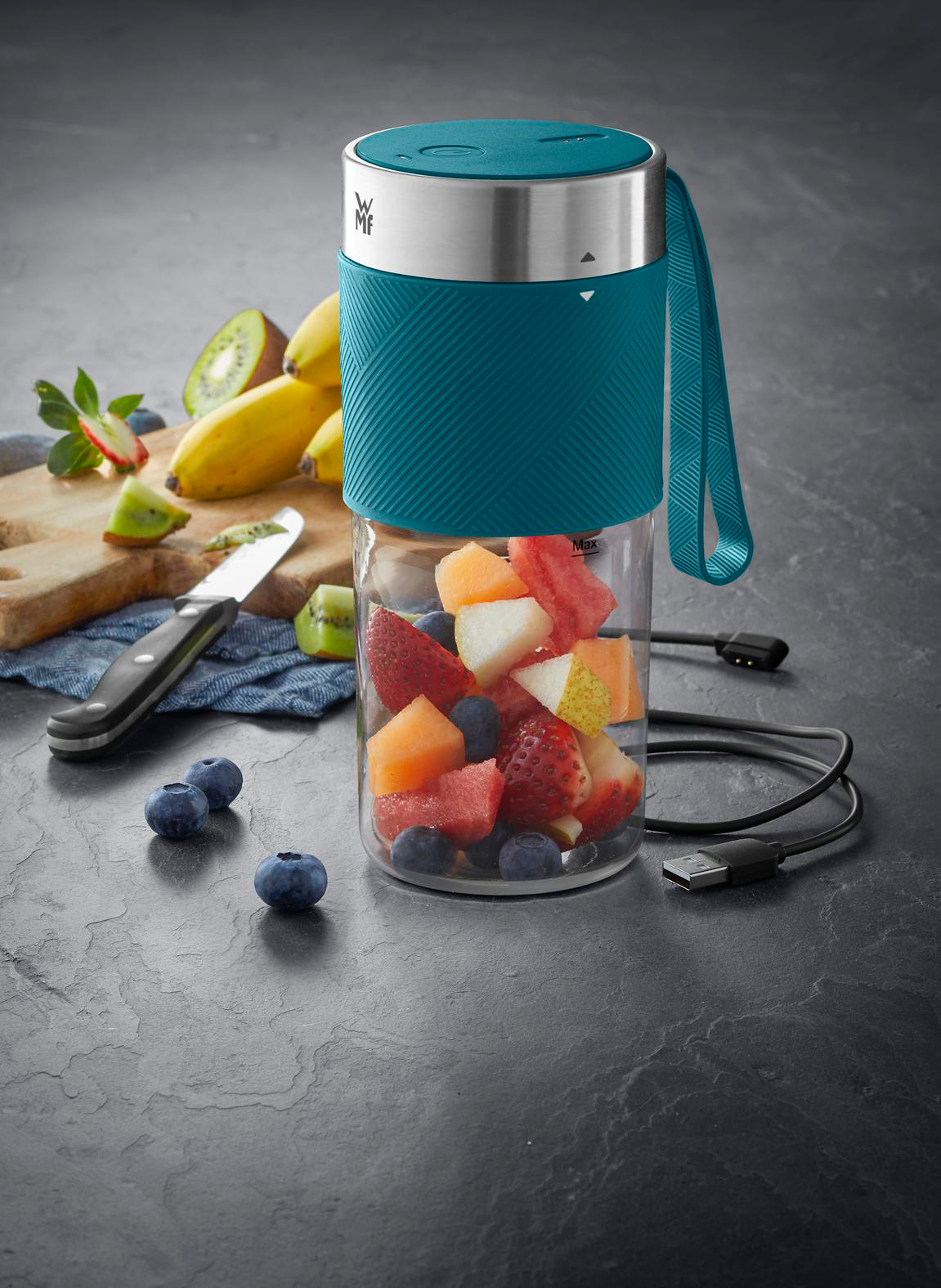 Ein Mixer mit Früchten, neben geschnittenen Früchten, Bananen und Blaubeeren. USB-Kabel vorhanden.