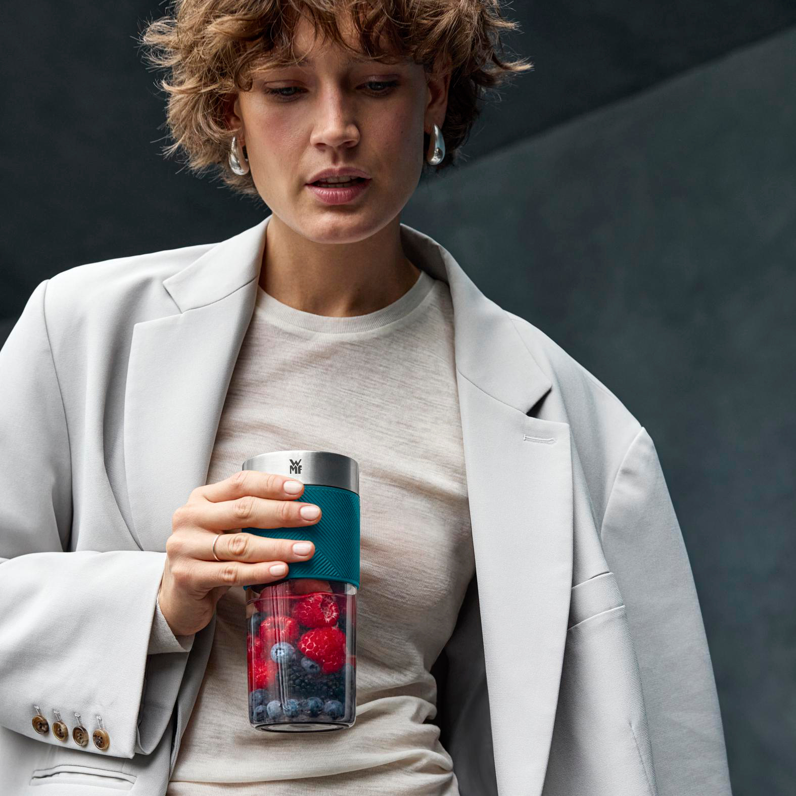 Eine Frau hält einen Smoothie-Becher mit Beeren. Sie trägt einen Blazer und Ohrringe. Der Hintergrund ist dunkelgrau.