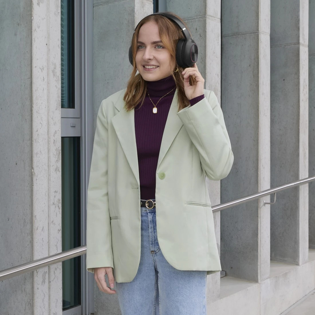 Una mujer con una chaqueta verde y auriculares sonríe al aire libre. Lleva un cuello alto morado, vaqueros y un collar.