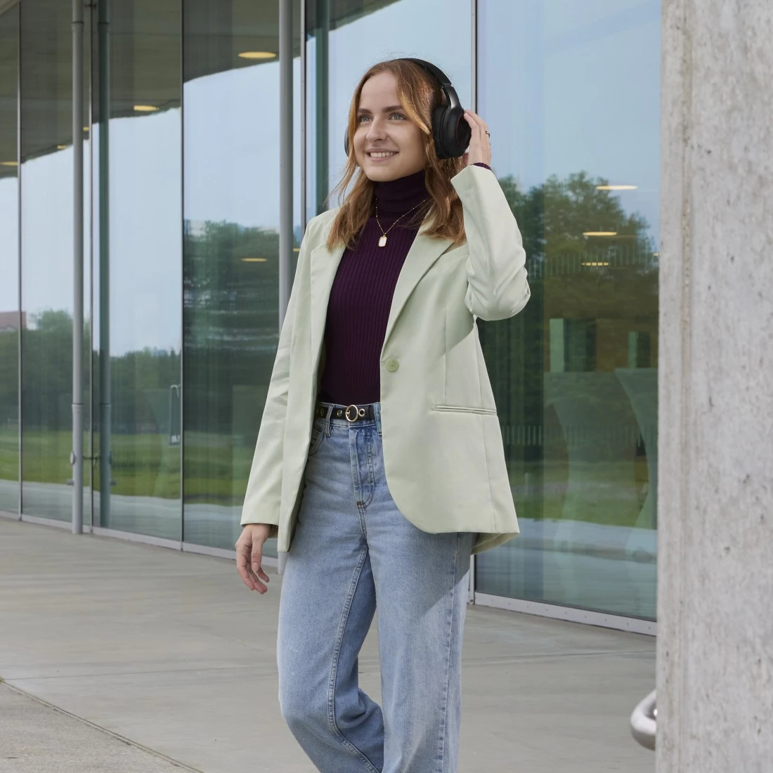 Mujer con auriculares, caminando, sonriendo. Lleva blazer verde, top morado, vaqueros. Fondo: edificio de cristal, hormigón.