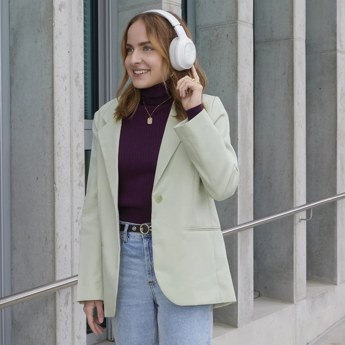 Una mujer con un cuello alto morado, una blazer verde claro y vaqueros, escuchando música con auriculares blancos, sonriendo.