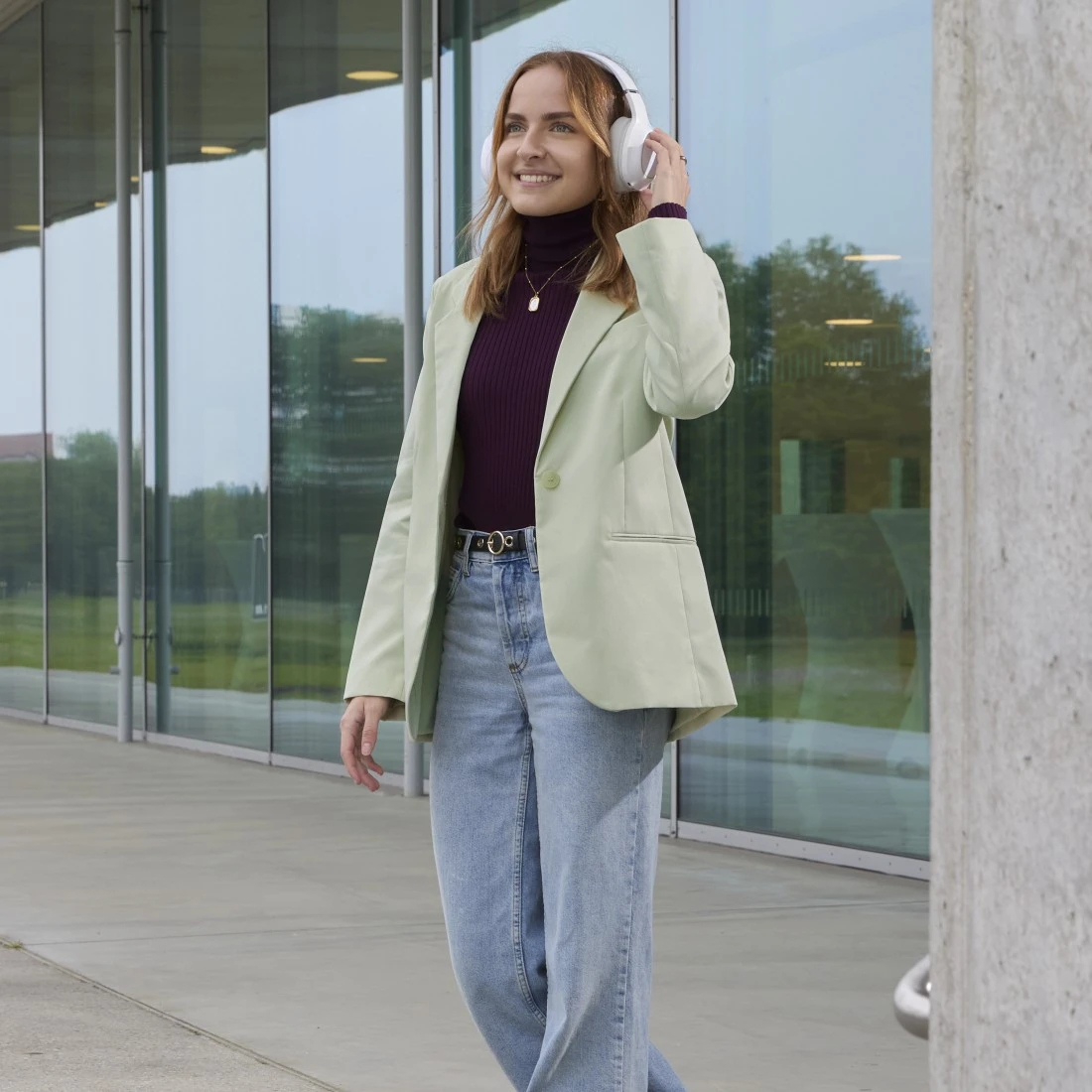 Mujer con auriculares, blazer y jeans, caminando cerca de un edificio de cristal. Ella está sonriendo. El fondo es un edificio.