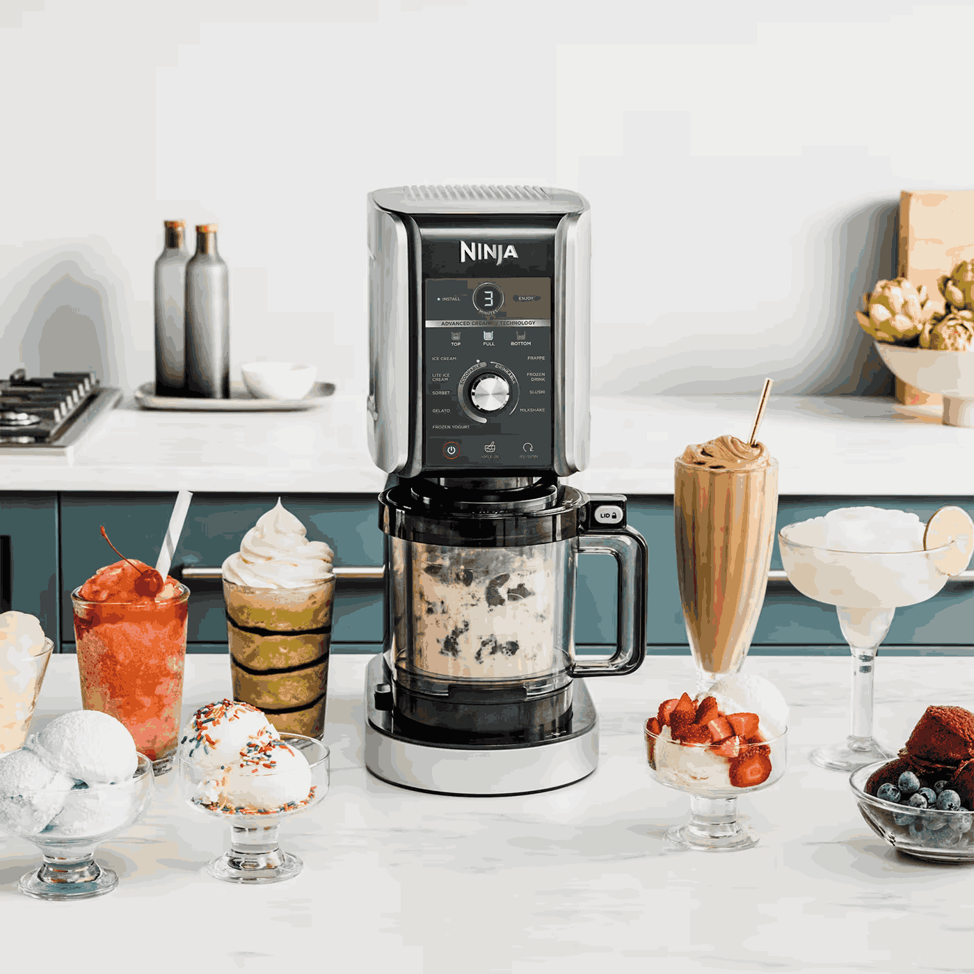 Una máquina Ninja Creami con varios postres: helado, sorbete y frappé, colocados sobre una encimera blanca. Se organizan diversos vasos y cuencos.