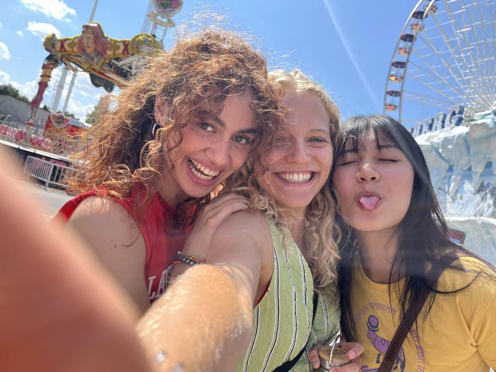 Drei lächelnde Frauen posieren für ein Selfie auf einem Jahrmarkt. Eine streckt die Zunge heraus. Im Hintergrund ein Riesenrad und ein Fahrgeschäft.
