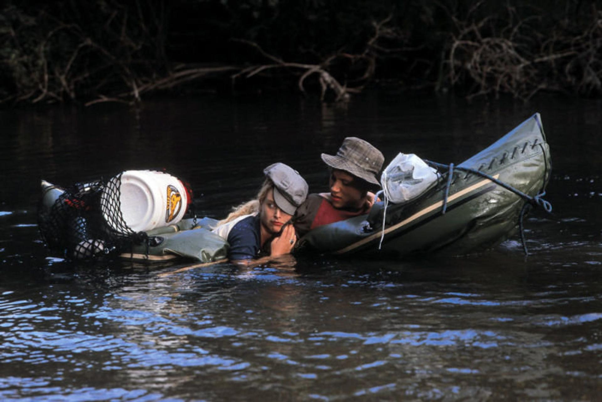 Zwei Personen in einem Floß auf einem Fluss. Eine Frau trägt eine Mütze, der Mann einen Hut. Ein weißer Eimer und andere Vorräte sind im Floß.